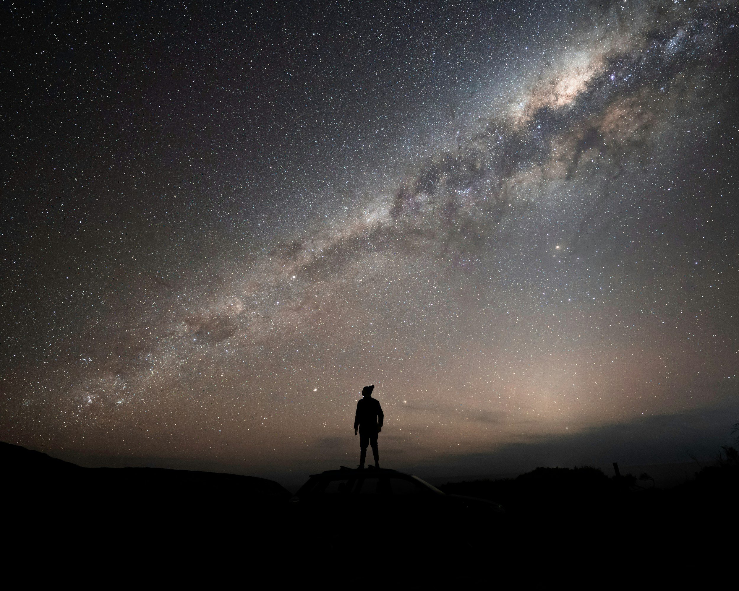 A person standing on a rock in a dark landscape, gazing at a star-filled night sky and the Milky Way galaxy.