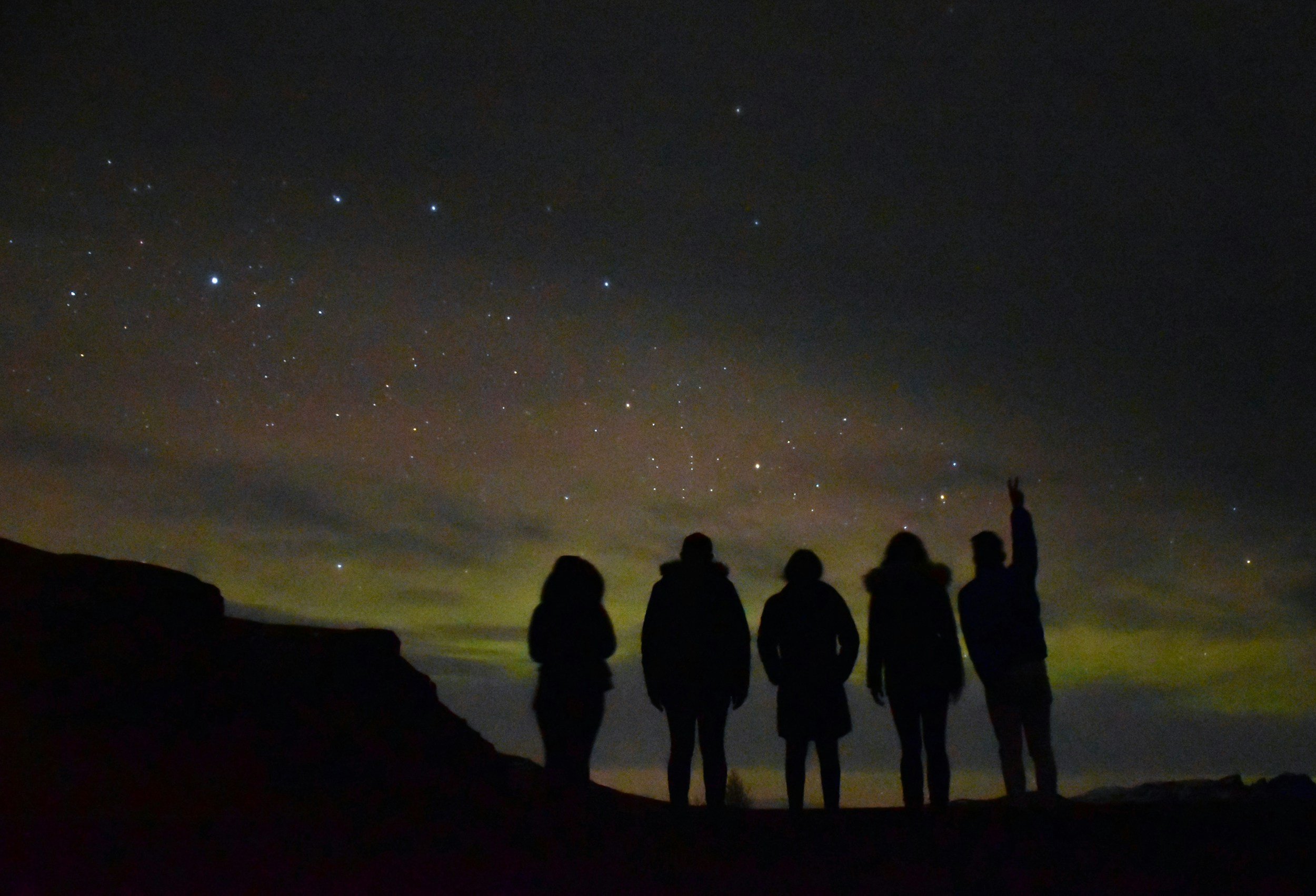 Five people standing outdoors at night looking at a starry sky, with some clouds and distant landscape visible.