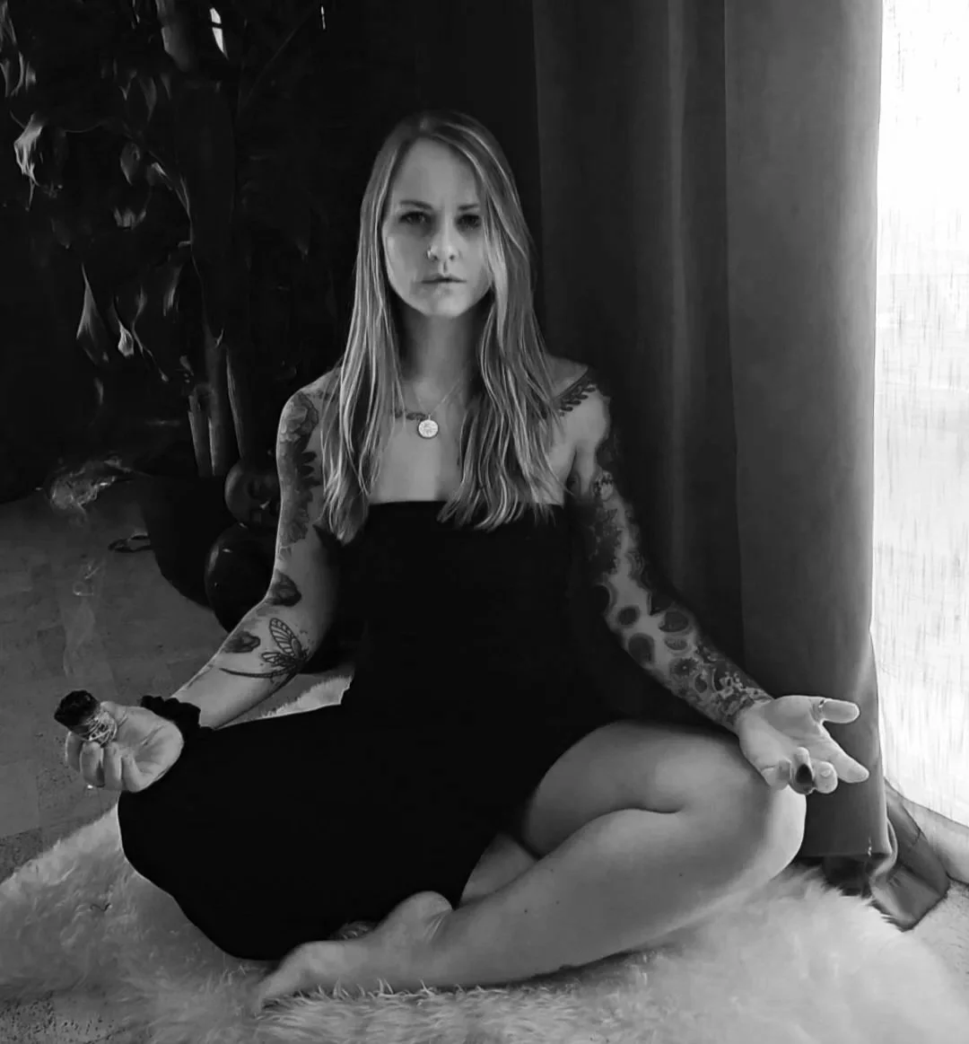 A woman with long hair and tattoos, sitting cross-legged on a fluffy rug in a room with a curtain and large plant, in a black and white photo, holding a small object.