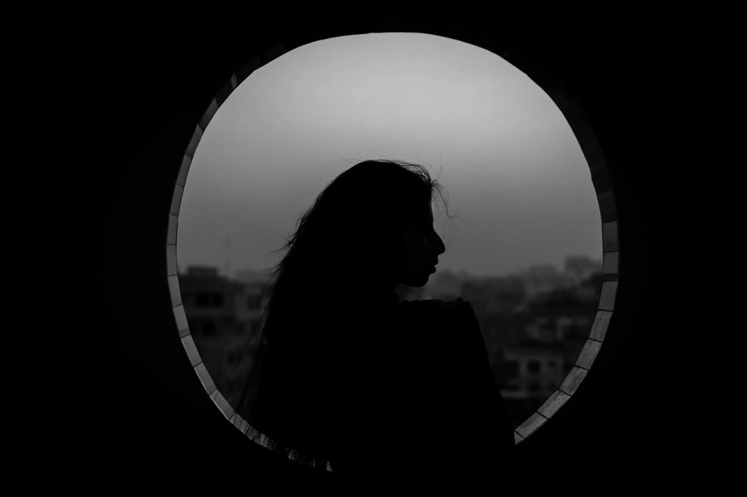Silhouette of a woman with long hair looking downward through a circular opening, cityscape background, black and white.