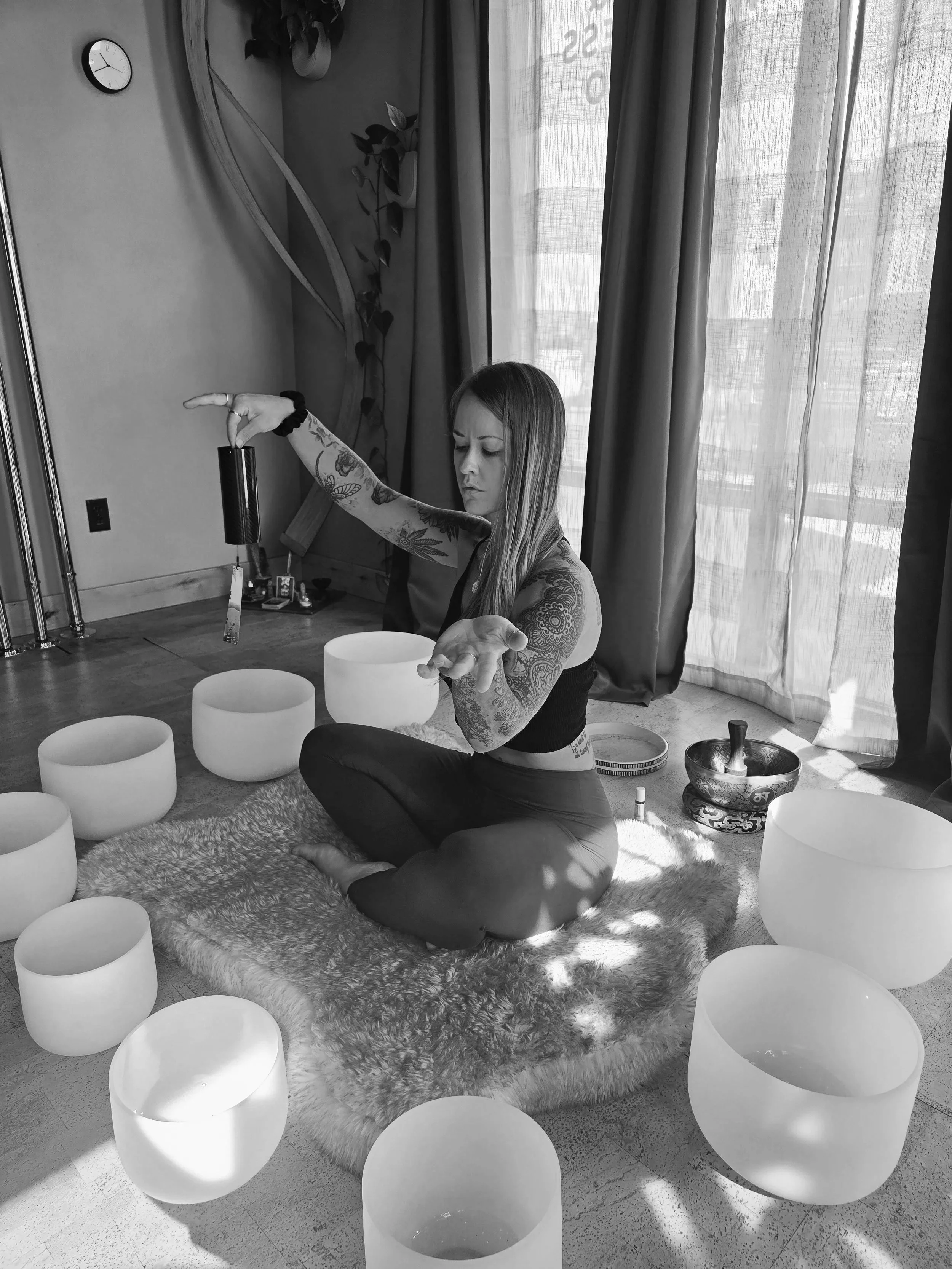 A woman with tattoos on her arms and a tattoo on her side, sitting cross-legged on a fluffy rug, playing crystal singing bowls in a room lit by natural light through large windows with curtains. There are several crystal bowls around her and a singing bowl in front of her.