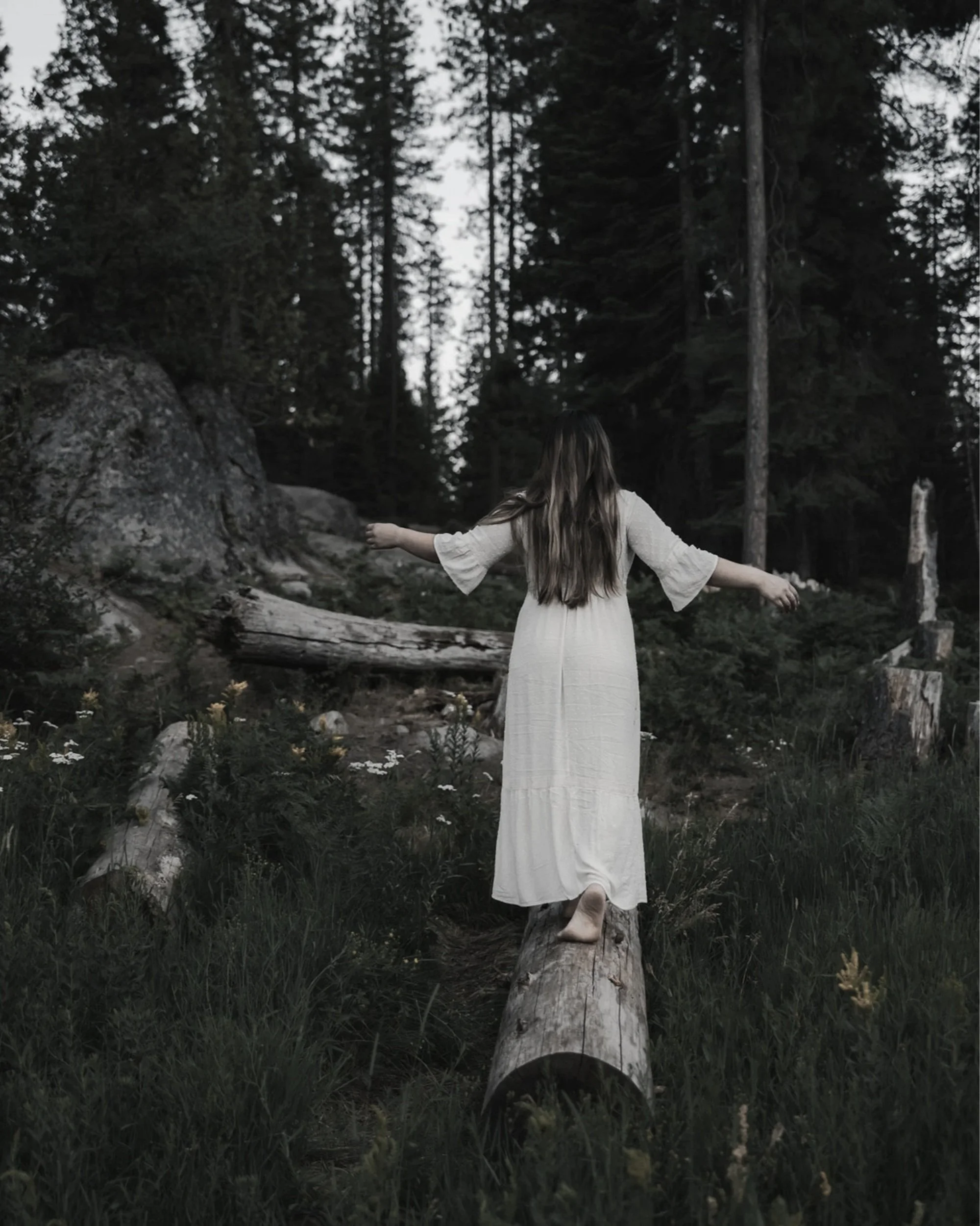 A woman with long hair, wearing a white dress, balancing on a fallen log in a dense forest with tall trees and rocks.