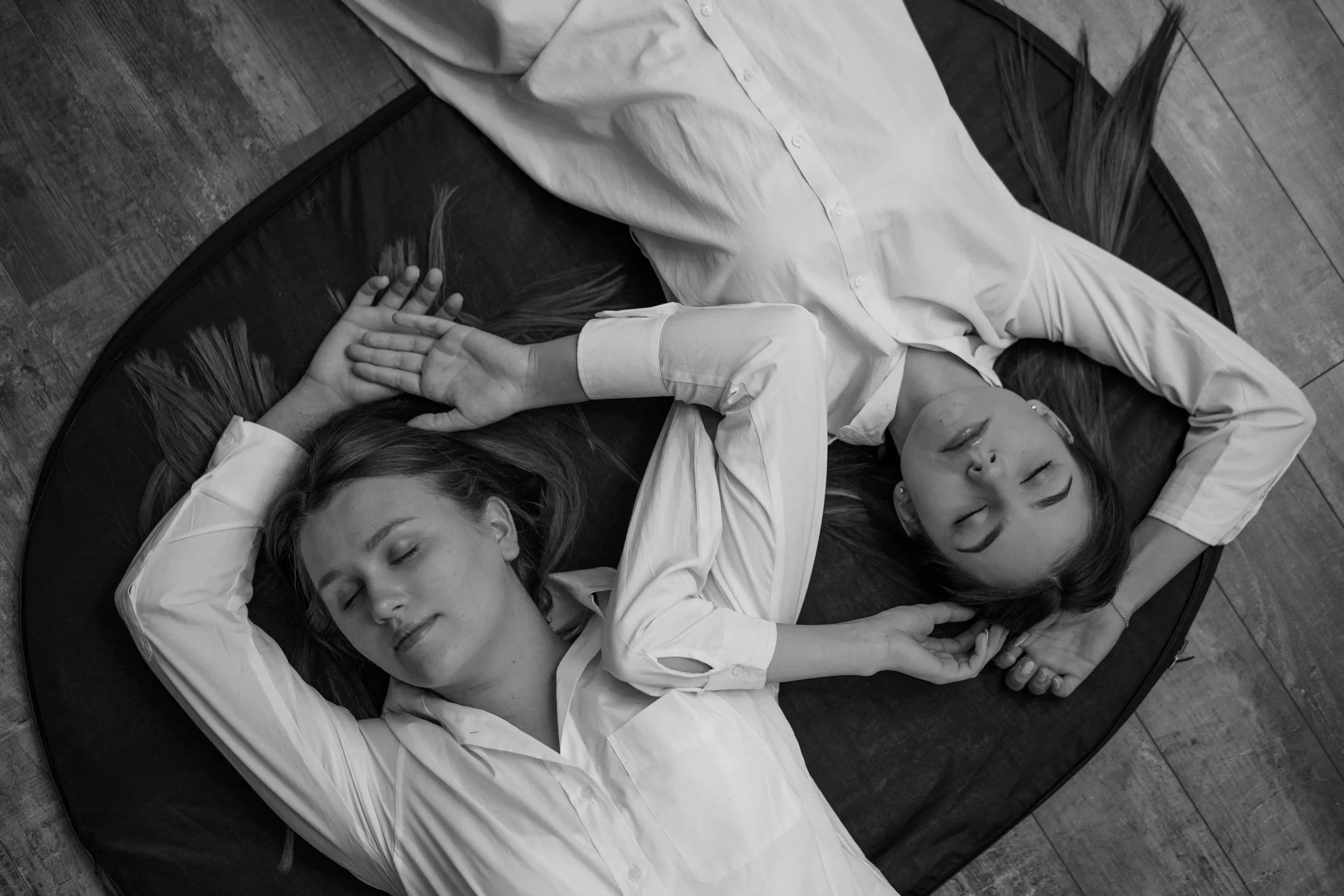 Two young women with long hair and light-colored shirts lie on their sides on a round black mat, with their eyes closed, on a wooden floor, holding hands.