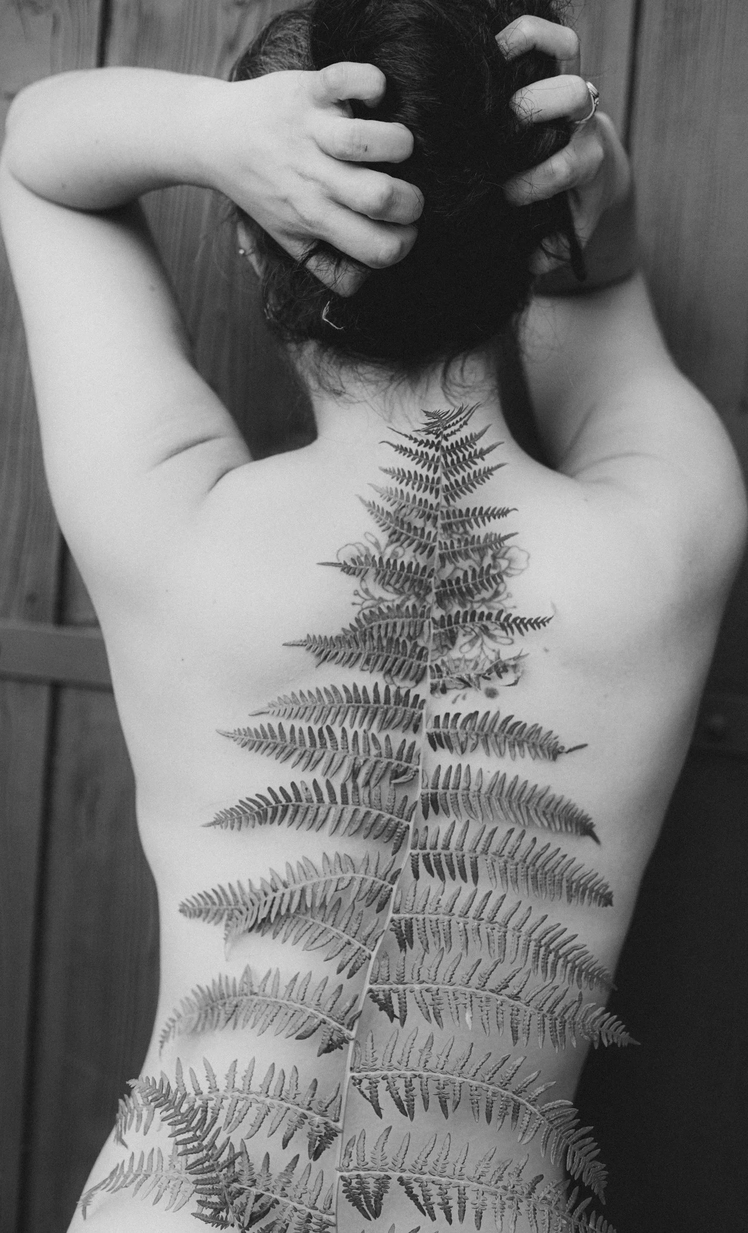 Black and white photograph of a person’s back with a large fern tattoo running down the spine. Hands are placed on the head, fingers in the hair, obscuring the face. The person appears to be topless, and the background is a wooden wall.
