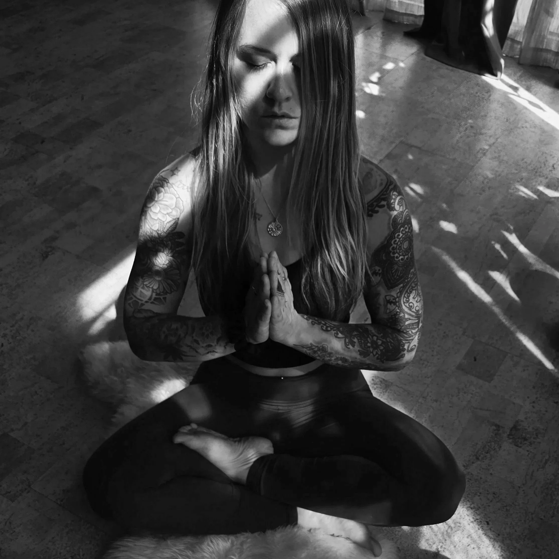 Black and white photo of a woman with long hair and tattoos on her arms, sitting on a rug, with her hands pressed together in a prayer position, eyes closed.