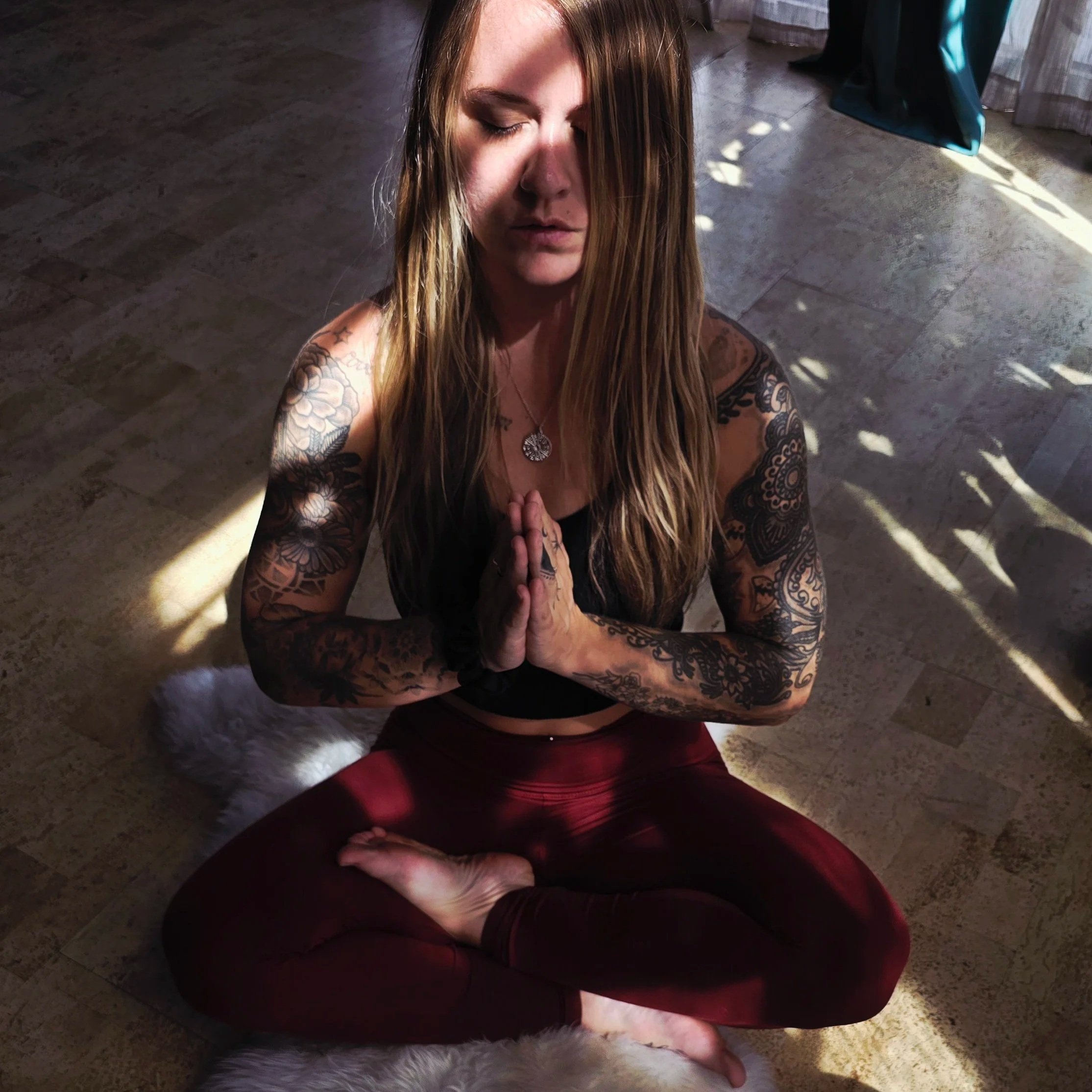 A woman with long hair and tattoos on her arms practicing yoga in a seated meditation pose with hands together in front of her chest, eyes closed, indoors near a window with sunlight.