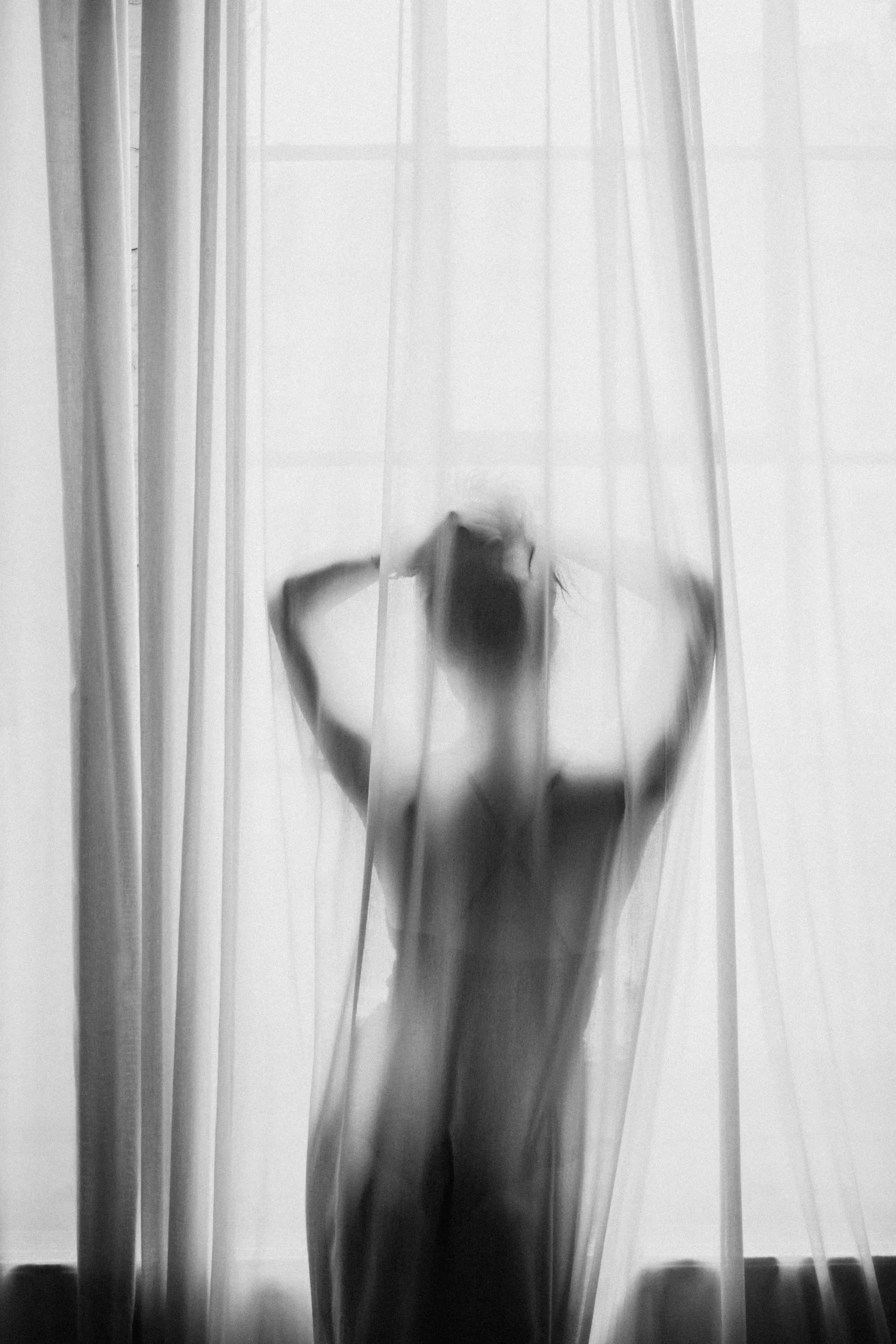 Black and white photo of a person standing behind sheer curtains, with their body and head blurred and obscured by the fabric.