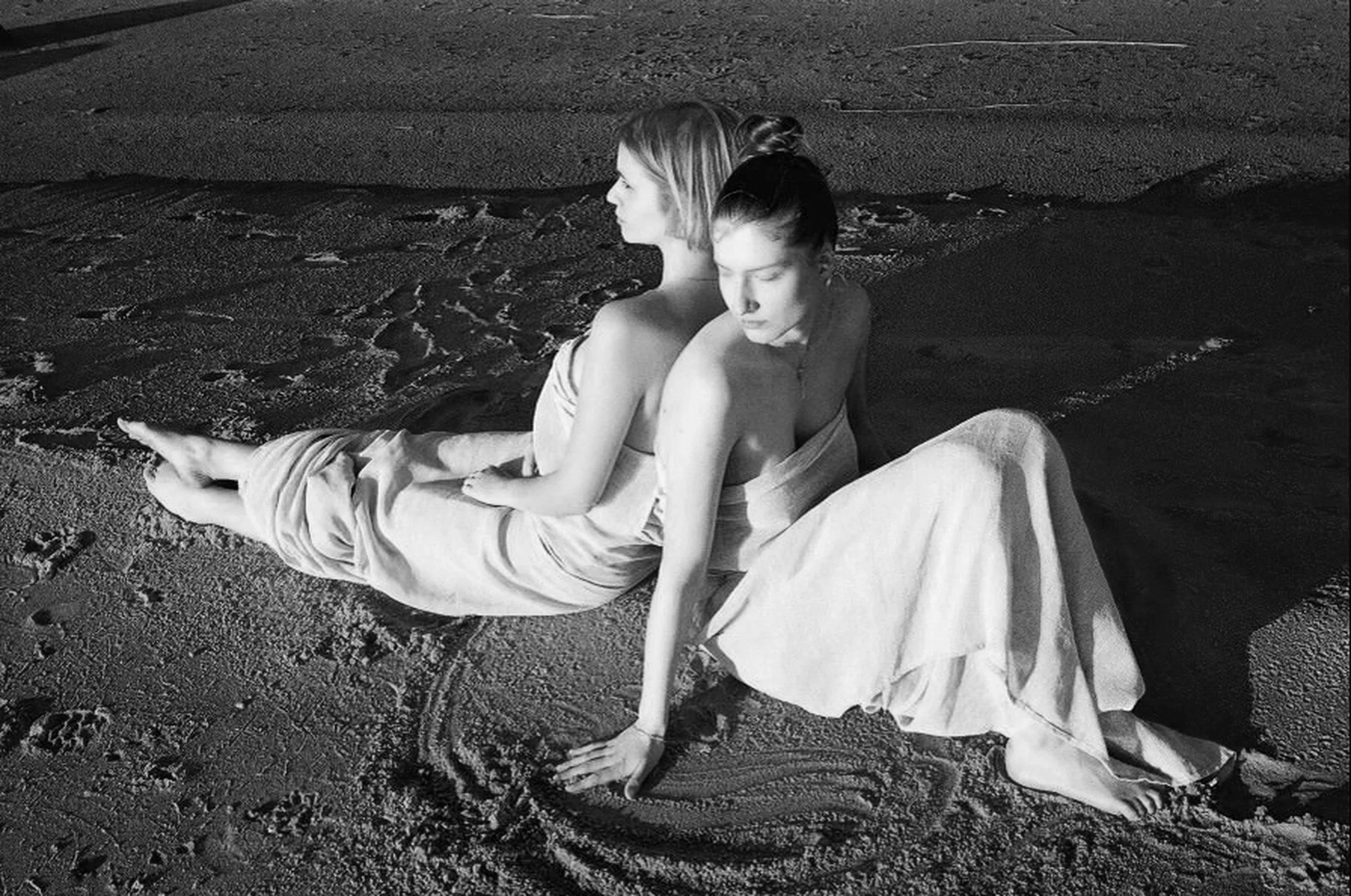 Black and white photo of two women sitting on the sand at the beach. One woman leans against the other with her eyes closed, while the other looks away. Both are wrapped in towels, with a beach chair nearby.