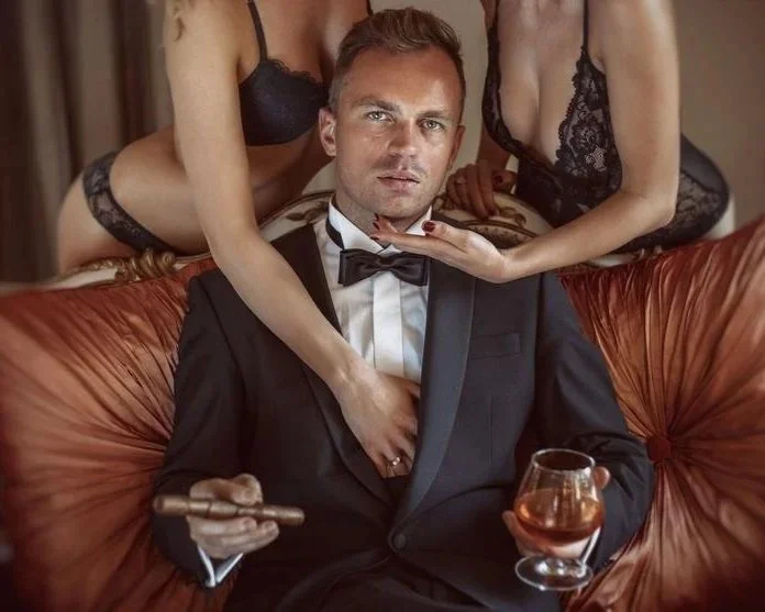 A man in a tuxedo sitting on a sofa with two women in lingerie, one on each side, one holding his chin and the other placing her hand on his chest, with one woman holding a cigar and the man holding a glass of liquor.