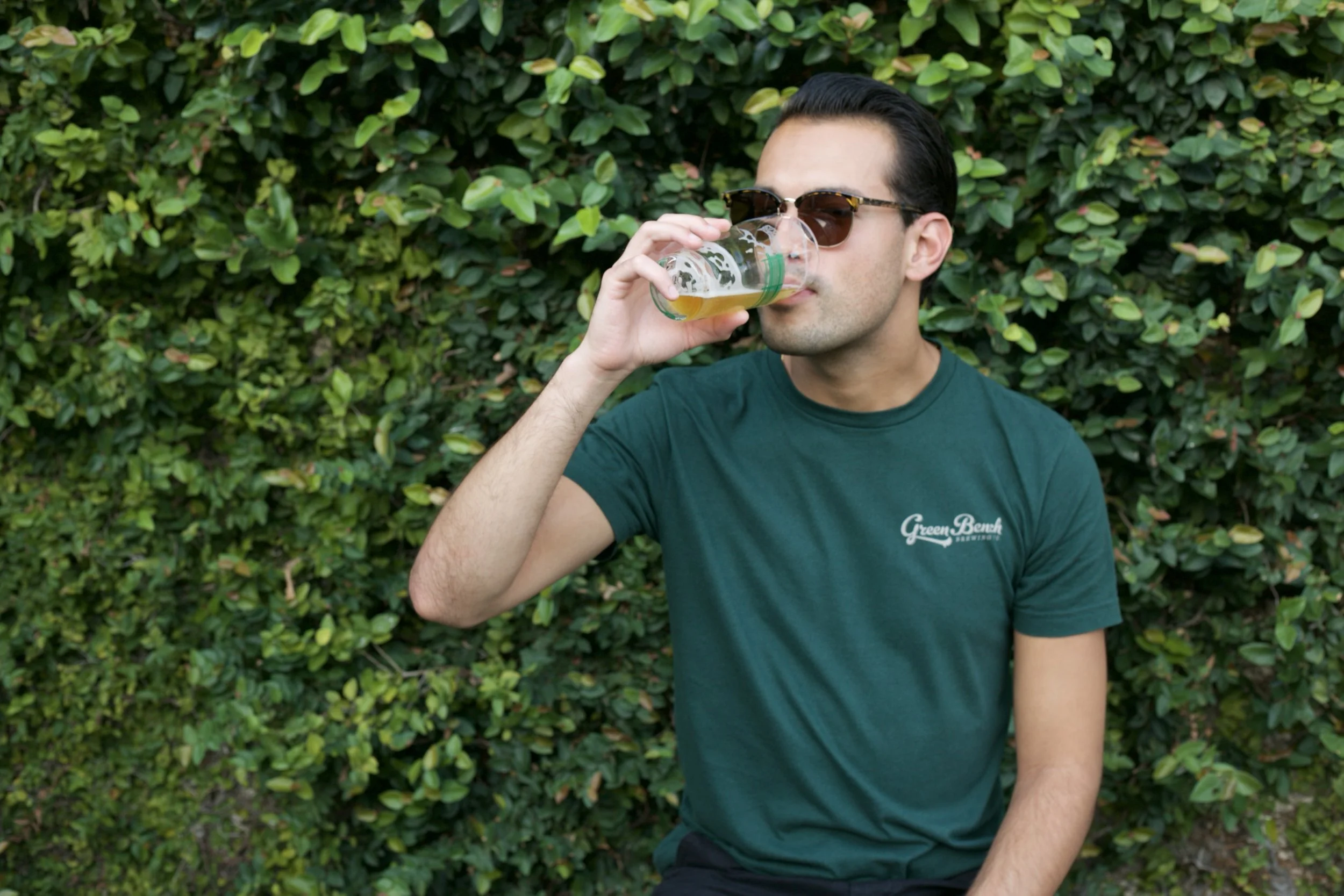 A man wearing sunglasses and a green T-shirt with 'Green Bench' on it, sitting outdoors and drinking from a glass of beer or cider, with green foliage behind him.