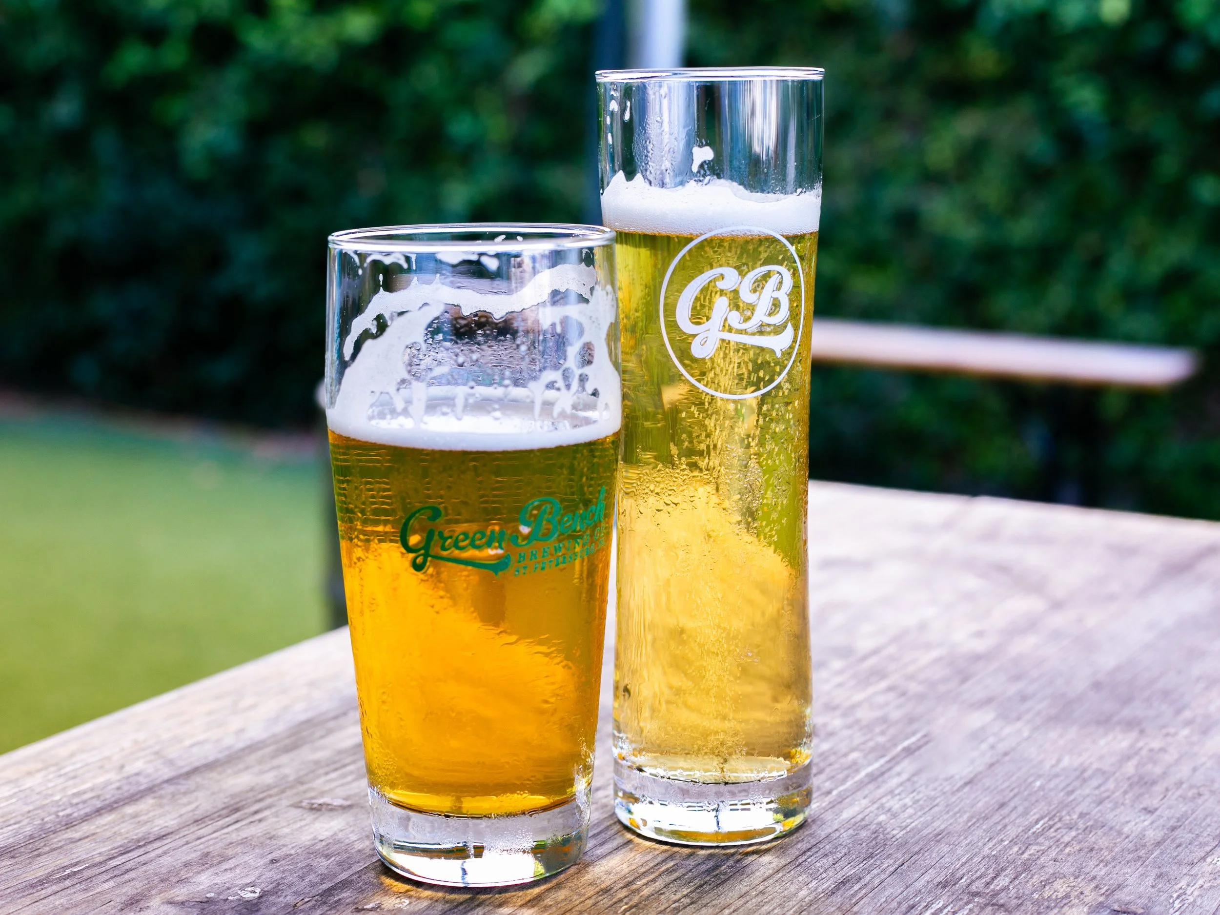 Two glasses of beer on a wooden table outdoors, one shorter glass labeled 'Green Bench' and one taller glass labeled 'G.B', with a blurred green background.