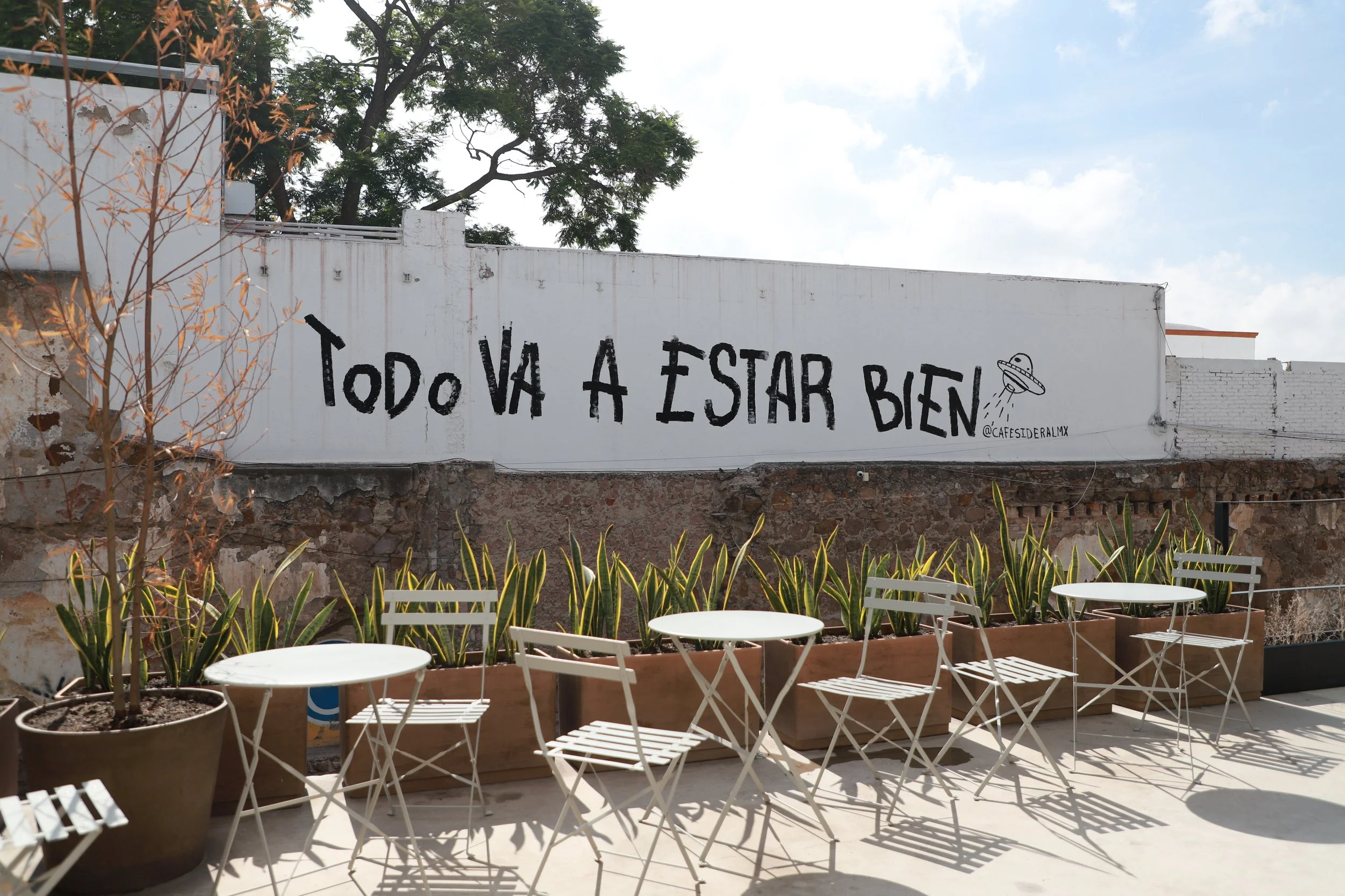 Outdoor seating area with white tables and chairs in front of a wall with graffiti that says 'Todo va a estar bien' and a drawing of a flying saucer. Potted plants are lined along the wall, with a brick wall and trees in the background.
