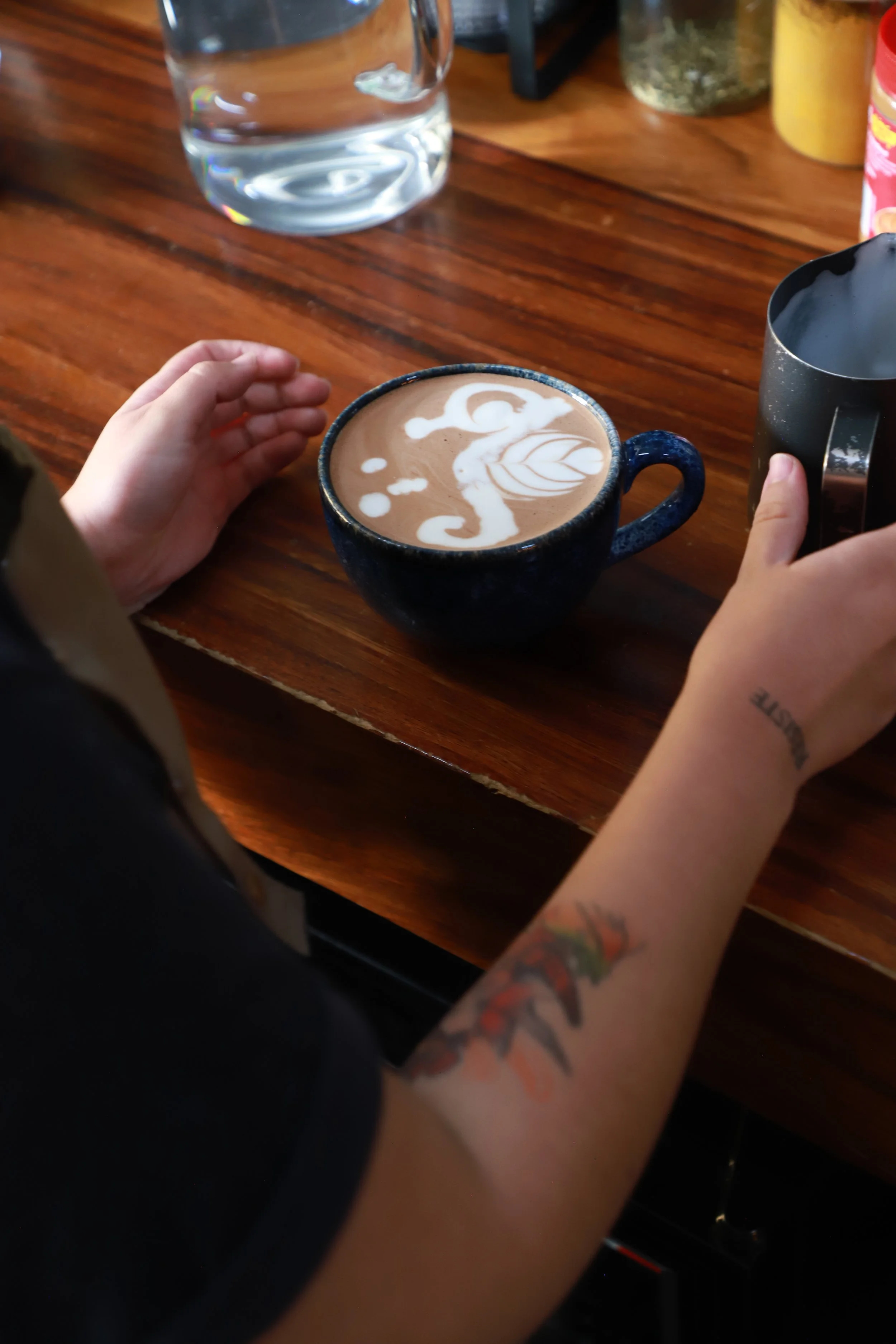 A person with a arm tattoo and a tattoo on their wrist prepares to add milk to a cup of coffee with latte art, on a wooden table.