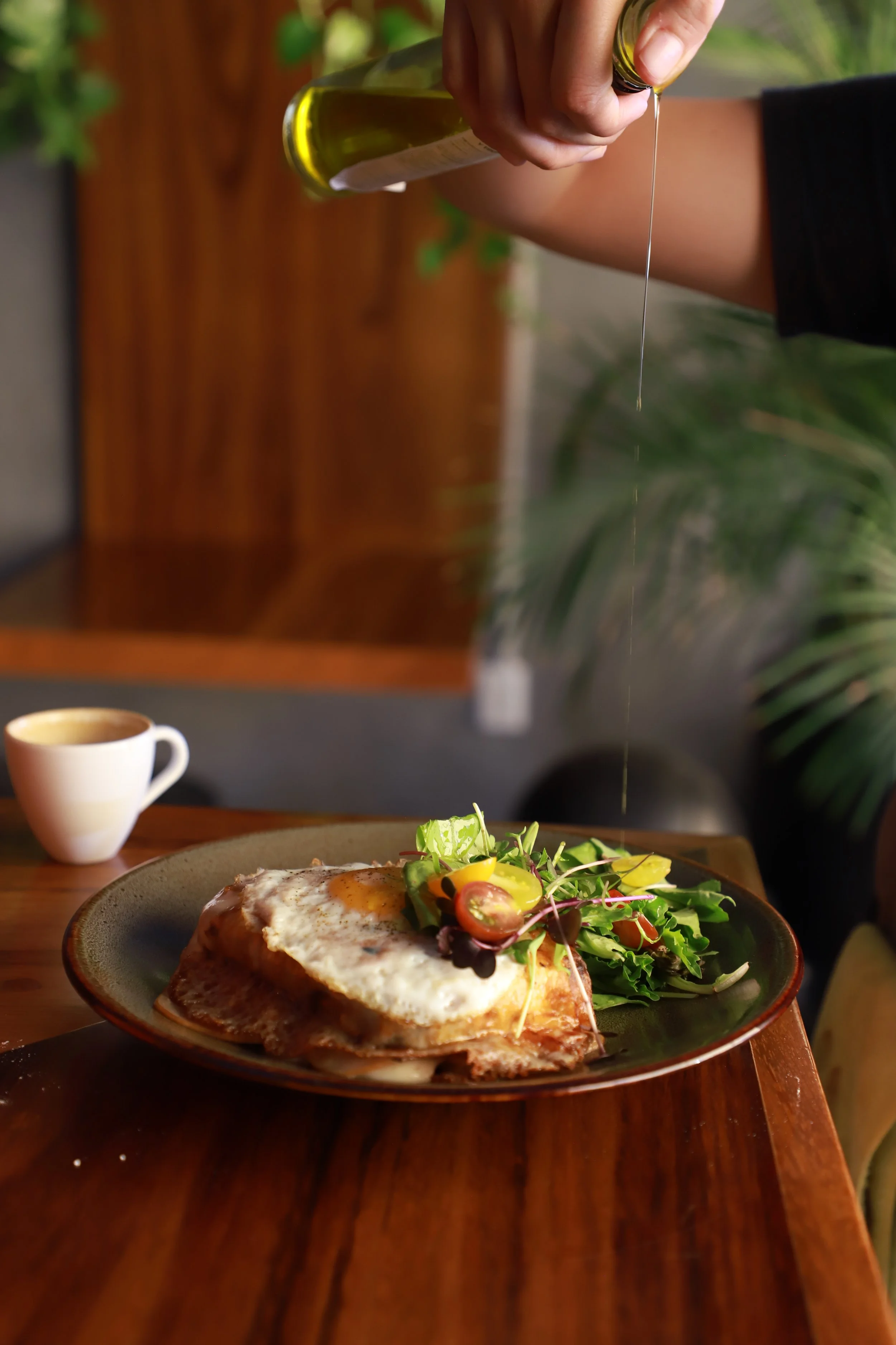 A hand pouring olive oil over a plate with a fried egg, salad, and toast. A cup of coffee is in the background.