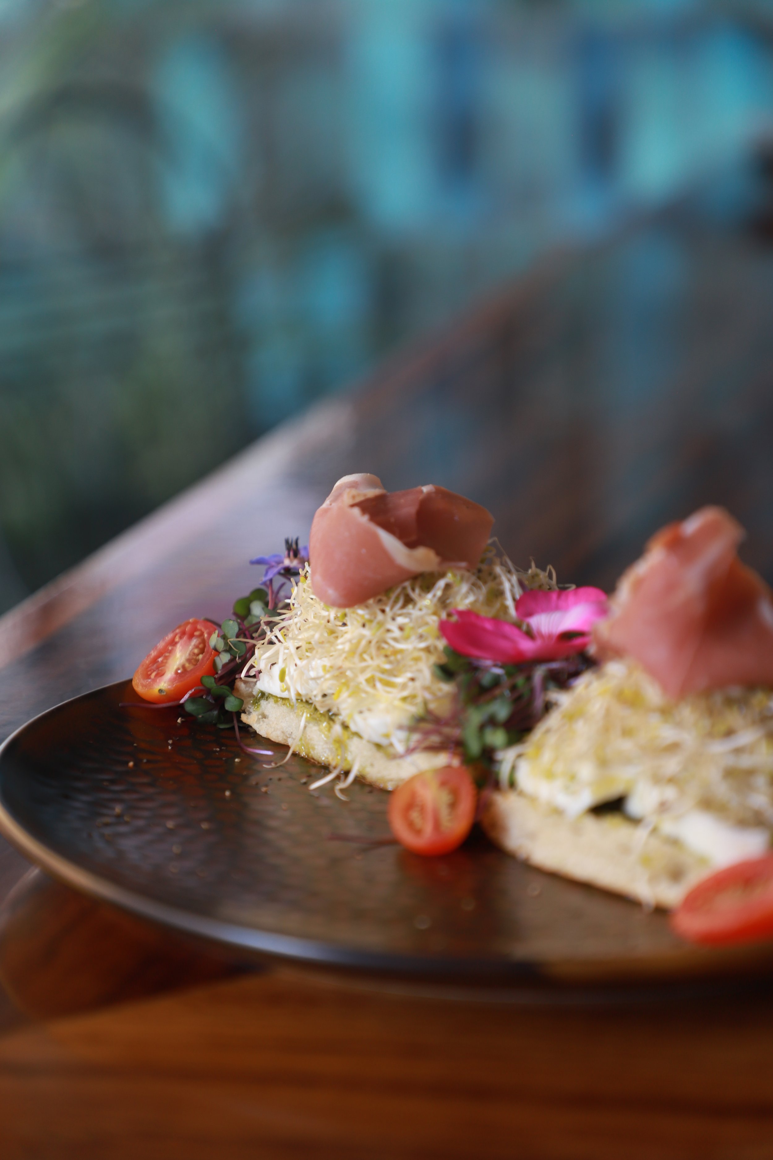 Slices of bread topped with shredded cheese, cherry tomatoes, microgreens, sliced ham, and edible flowers on a black textured plate.