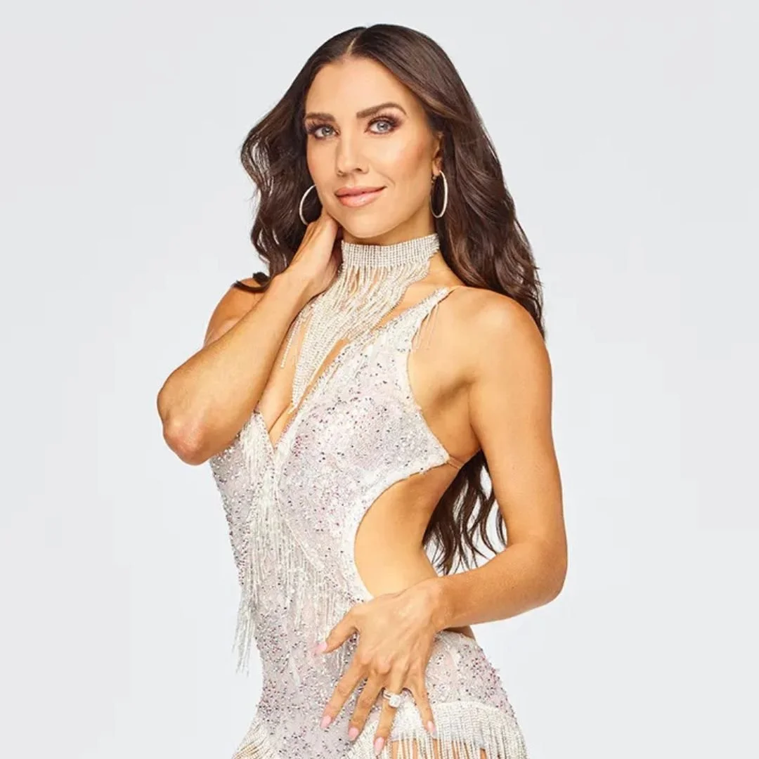 A woman with long, dark hair wearing a sparkly, cut-out, beige dress with silver jewelry including hoop earrings and a choker, posing with her hand on her face and hips.