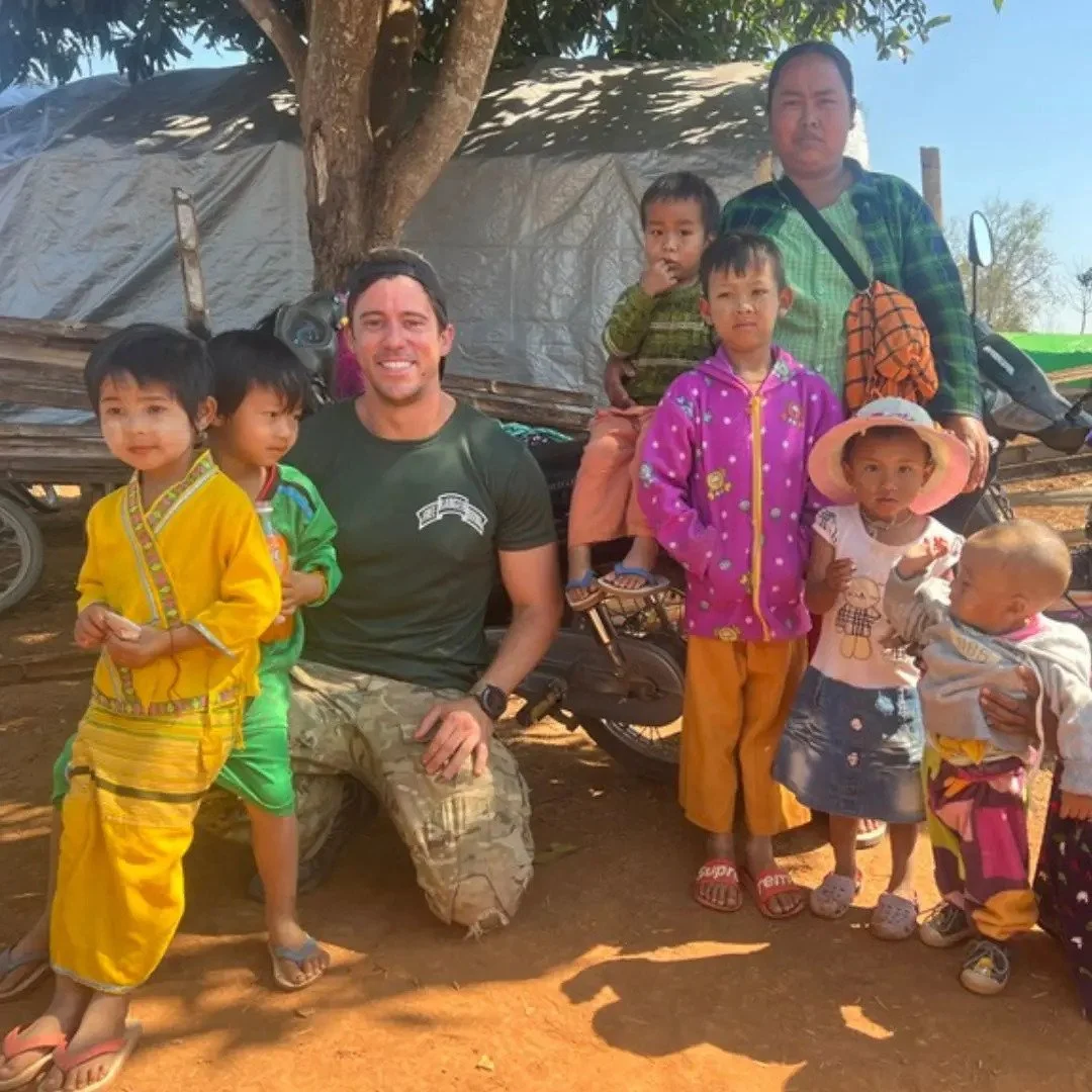 A group of children and two adults posing outdoors under a tree. The children are wearing colorful clothes, and the adults are smiling. In the background, there is a tent and some bicycles.