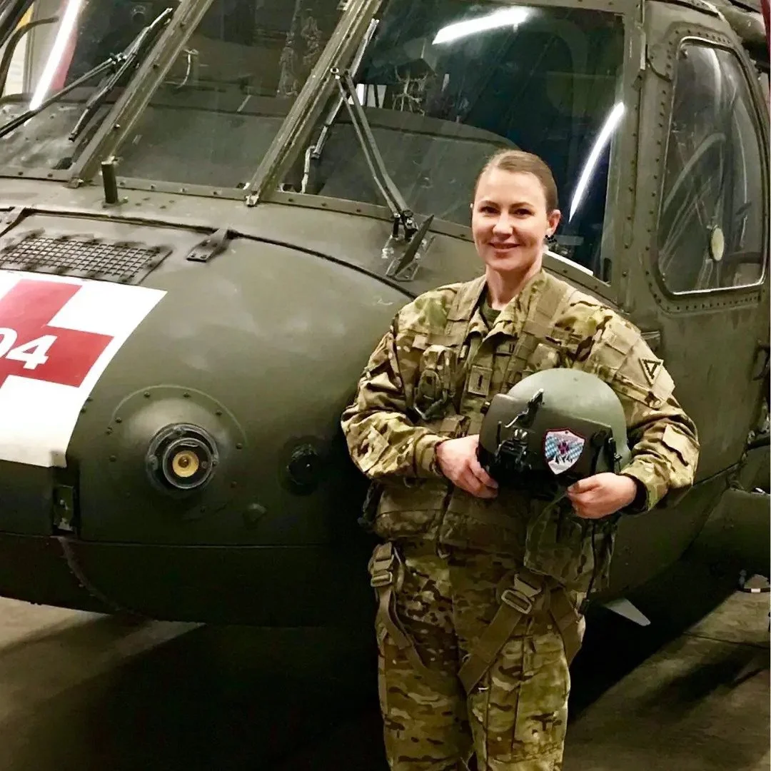 A woman in military uniform holding a helmet with a patch, standing next to a military helicopter with a red cross sign.
