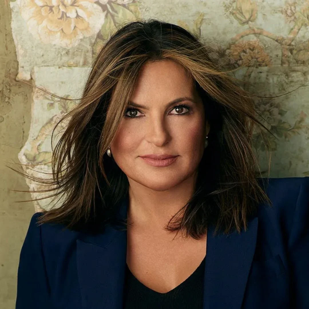 A woman with shoulder-length brown hair and light makeup, wearing pearl earrings and a navy blue blazer, looking directly at the camera. Background features a floral wall mural.