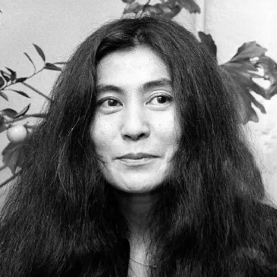 Black and white portrait of a woman with long, wavy hair, standing in front of leafy plants.