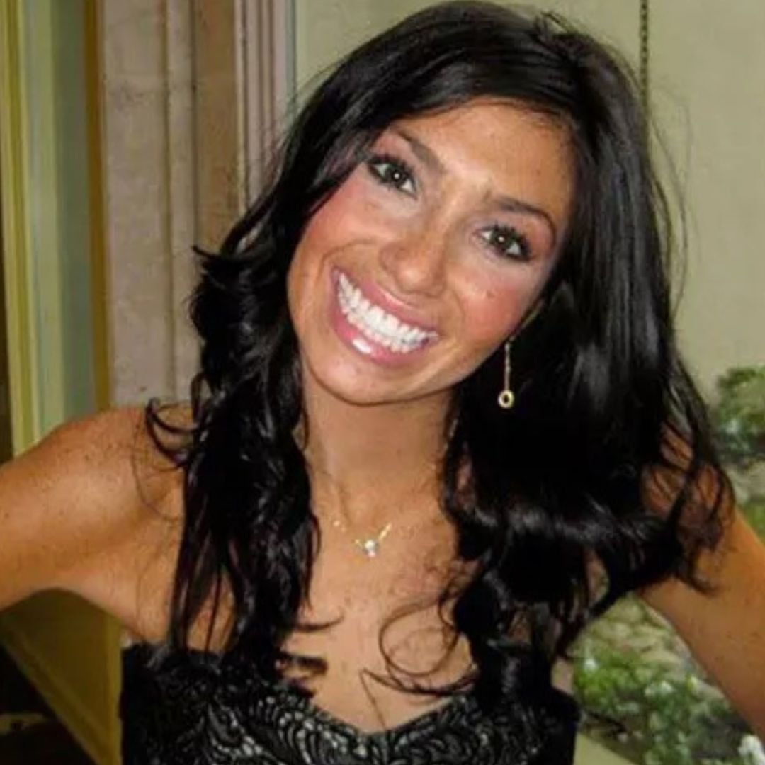 A woman with long black wavy hair smiling, wearing a black dress with lace details, earrings, a necklace, and makeup, in an indoor setting.