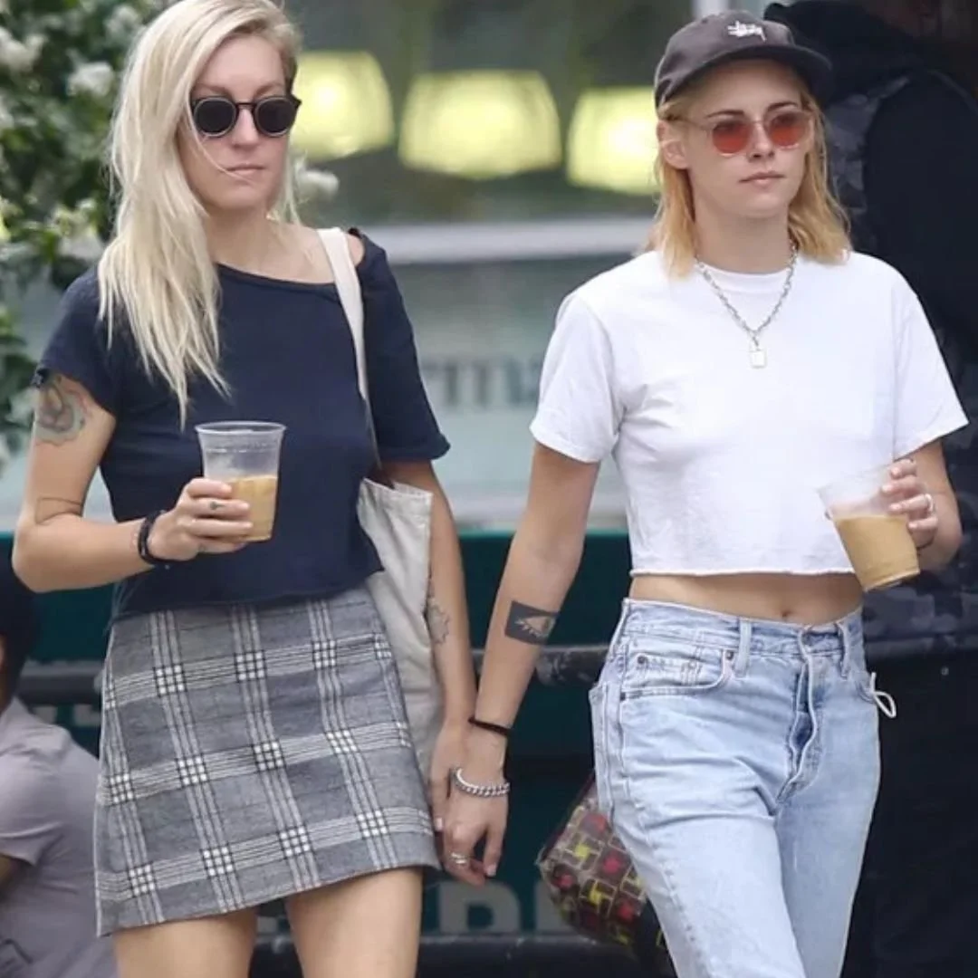 Two young women holding drinks and holding hands, walking outdoors. One woman has blonde hair, wearing a black t-shirt, plaid skirt, sunglasses, and a beige bag. The other woman has blonde hair, wearing a white crop top, light jeans, sunglasses, a cap, and a necklace.