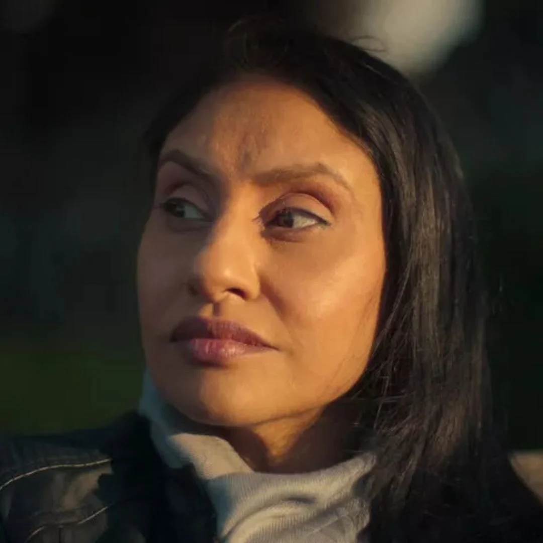 Close-up of a woman with dark hair, looking to the side with a contemplative expression, outdoors in dim lighting.