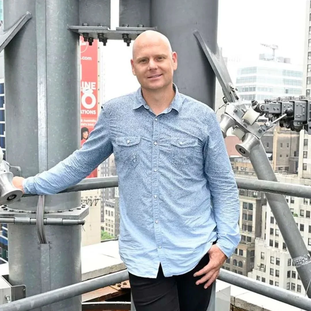 A man standing on a rooftop in a city, holding onto a metallic structure with buildings in the background.