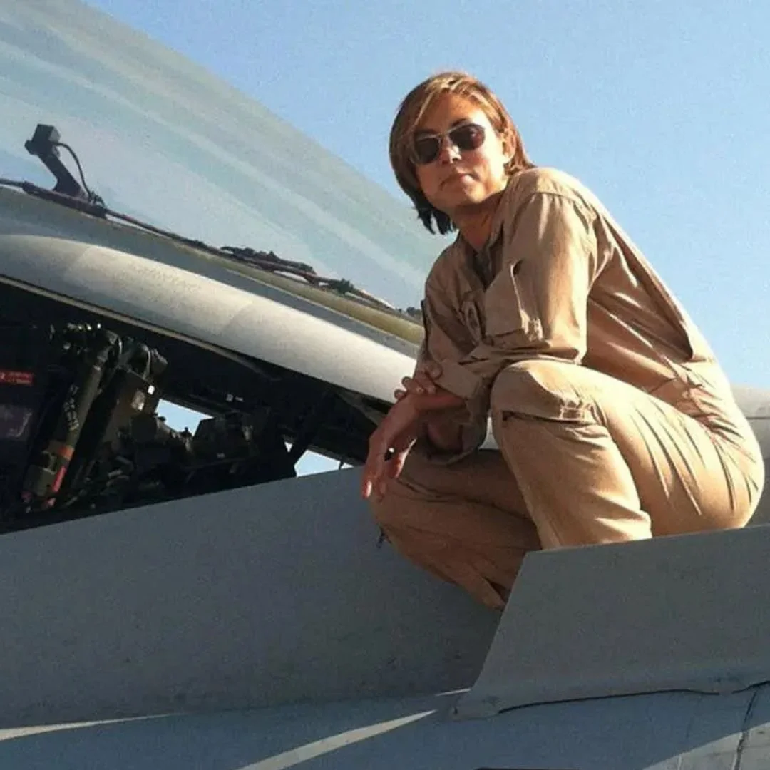 A woman with short hair, sunglasses, in a tan jumpsuit, crouching on the wing of a fighter jet, looking at the camera under a clear blue sky.