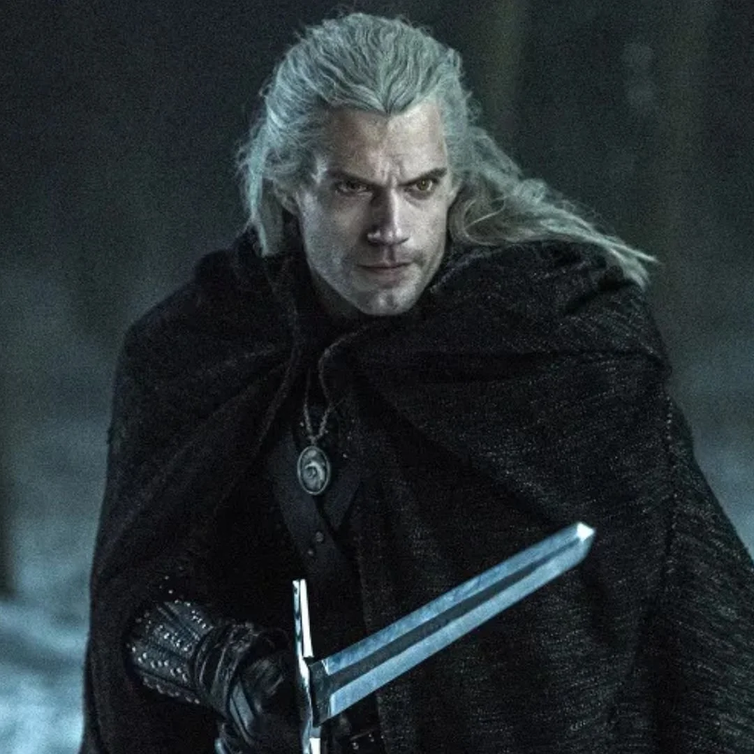 A character with long grey hair and a serious expression, dressed in dark clothing, holding a sword with a grey background.