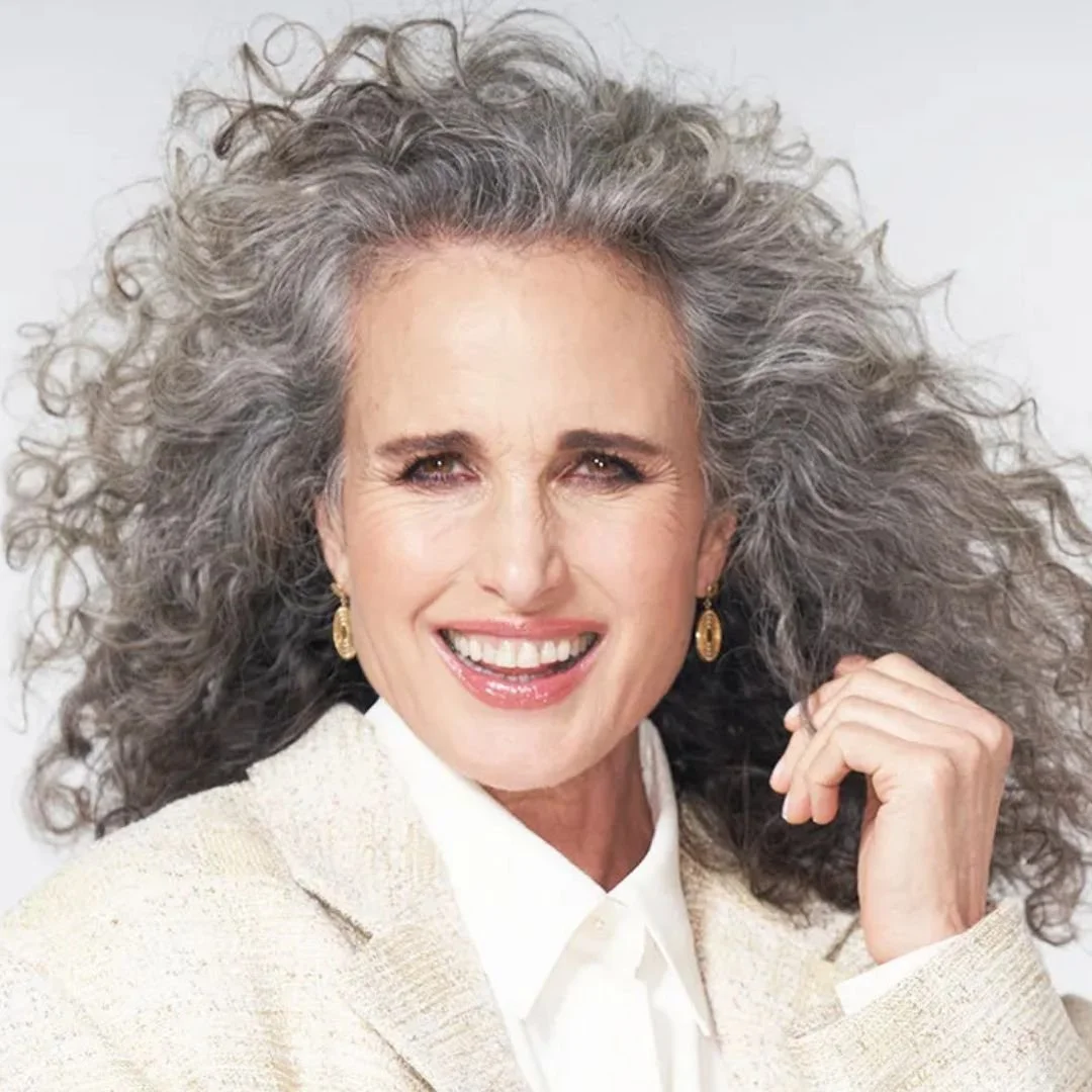 A smiling older woman with curly gray hair, wearing gold earrings, a white shirt, and a beige blazer, posing against a neutral background.