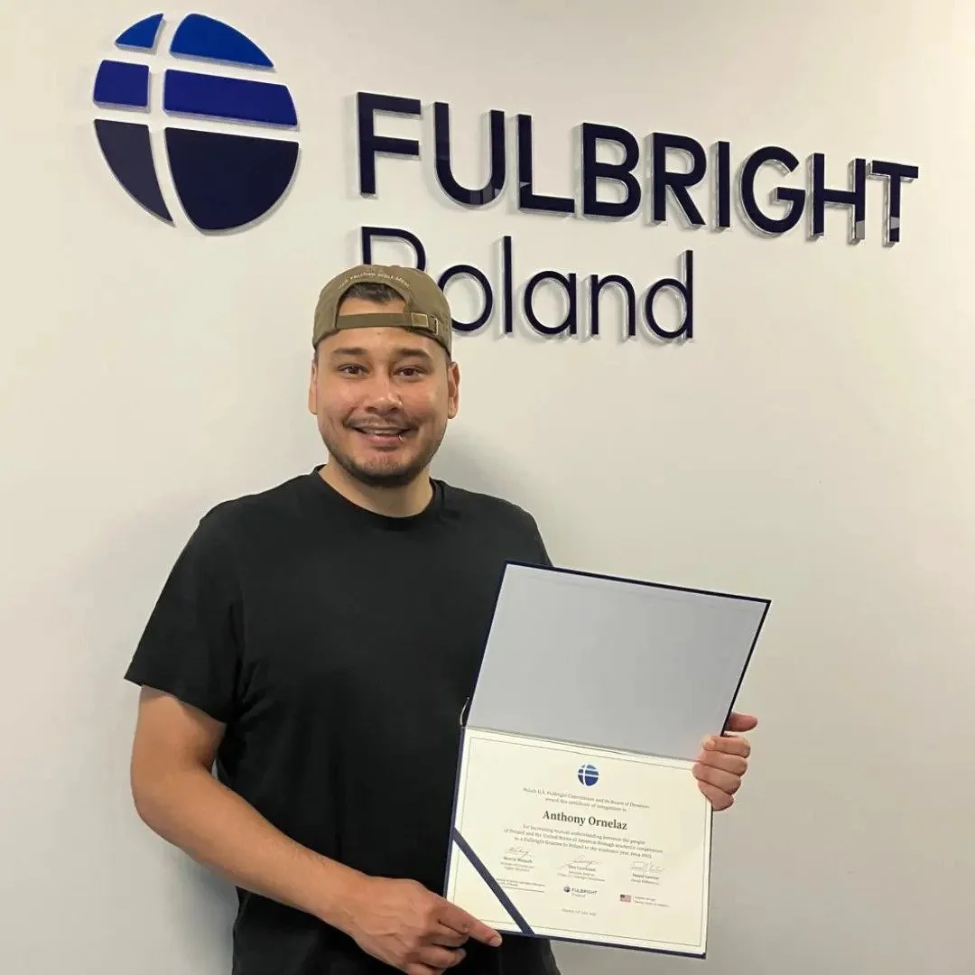 Man holding a diploma in front of a wall with the logos 'Fulbright' and 'Poland'.