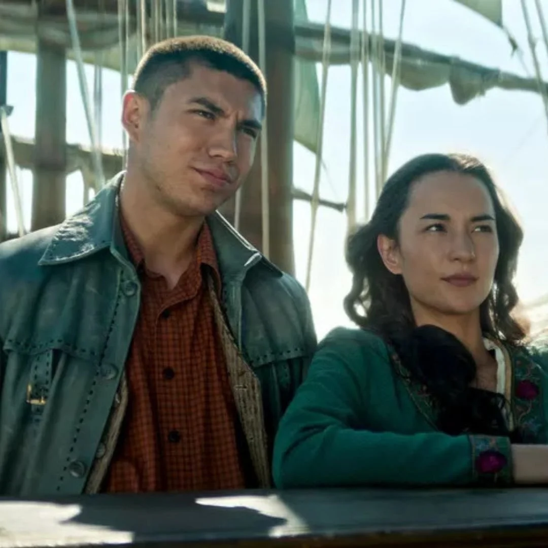 A man and woman standing outdoors in front of a structure with ropes, possibly a ship or dock, with the man squinting and the woman looking slightly to the side.