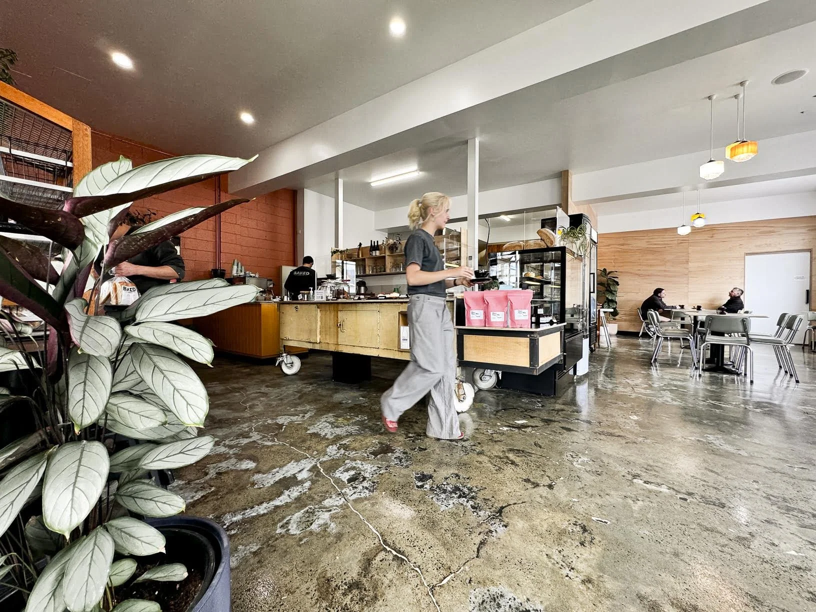 Interior of a modern coffee shop with a counter in the background, a barista serving customers, and two people sitting at a table on the right side. There are plants in the foreground and colorful hanging lights on the ceiling.