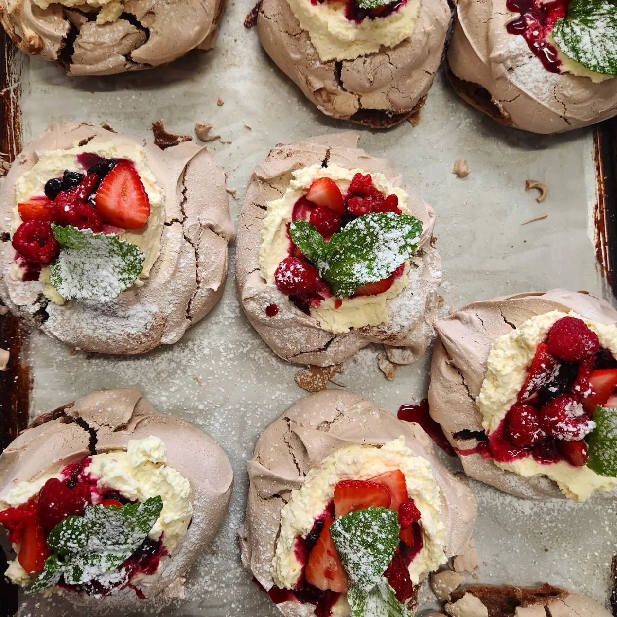 Meringue desserts topped with fresh strawberries, mixed berries, whipped cream, and mint leaves, dusted with powdered sugar.
