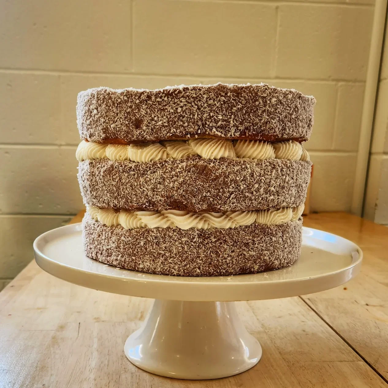 Four-layer cake with chocolate-rolled icing on each layer, stacked on a white cake stand on a wooden table against a beige brick wall.