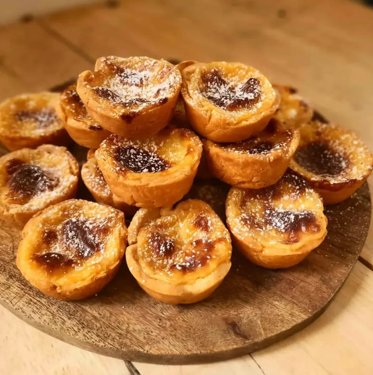 Close-up of a wooden board with several Portuguese custard tarts sprinkled with powdered sugar.