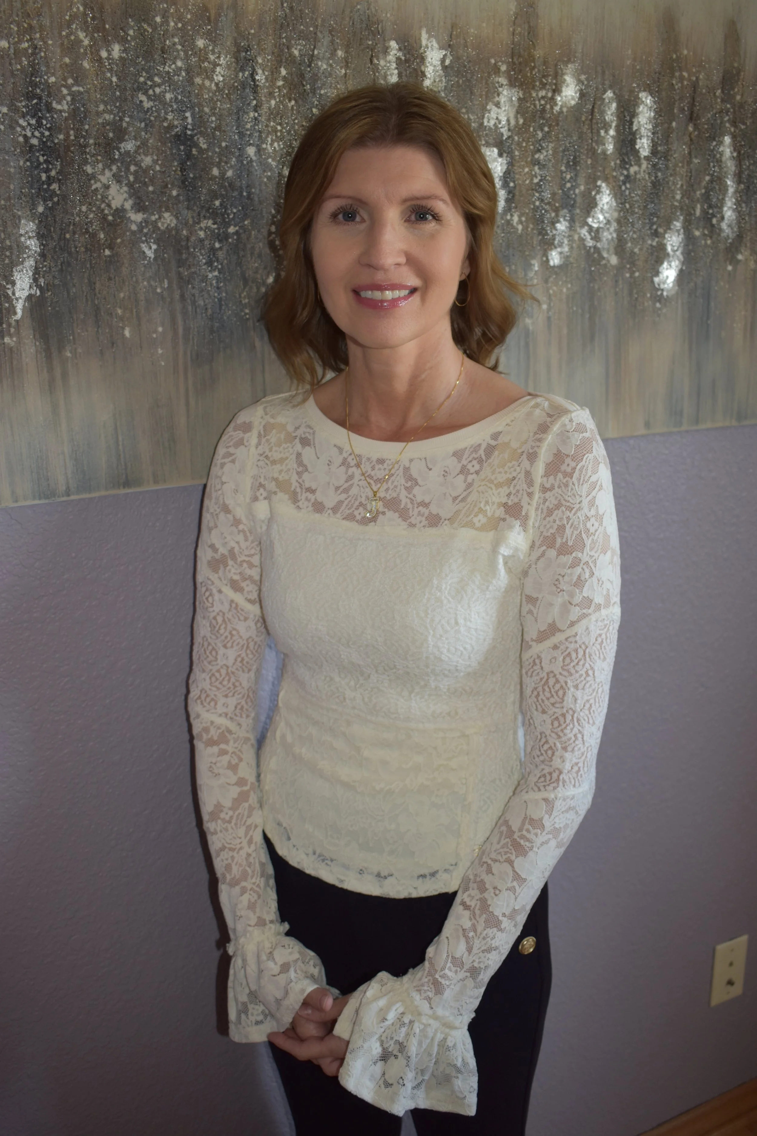 A woman with reddish-brown hair wearing a cream-colored lace long-sleeve top, black pants, jewelry including a necklace and earrings, standing against a textured wall with abstract paint.