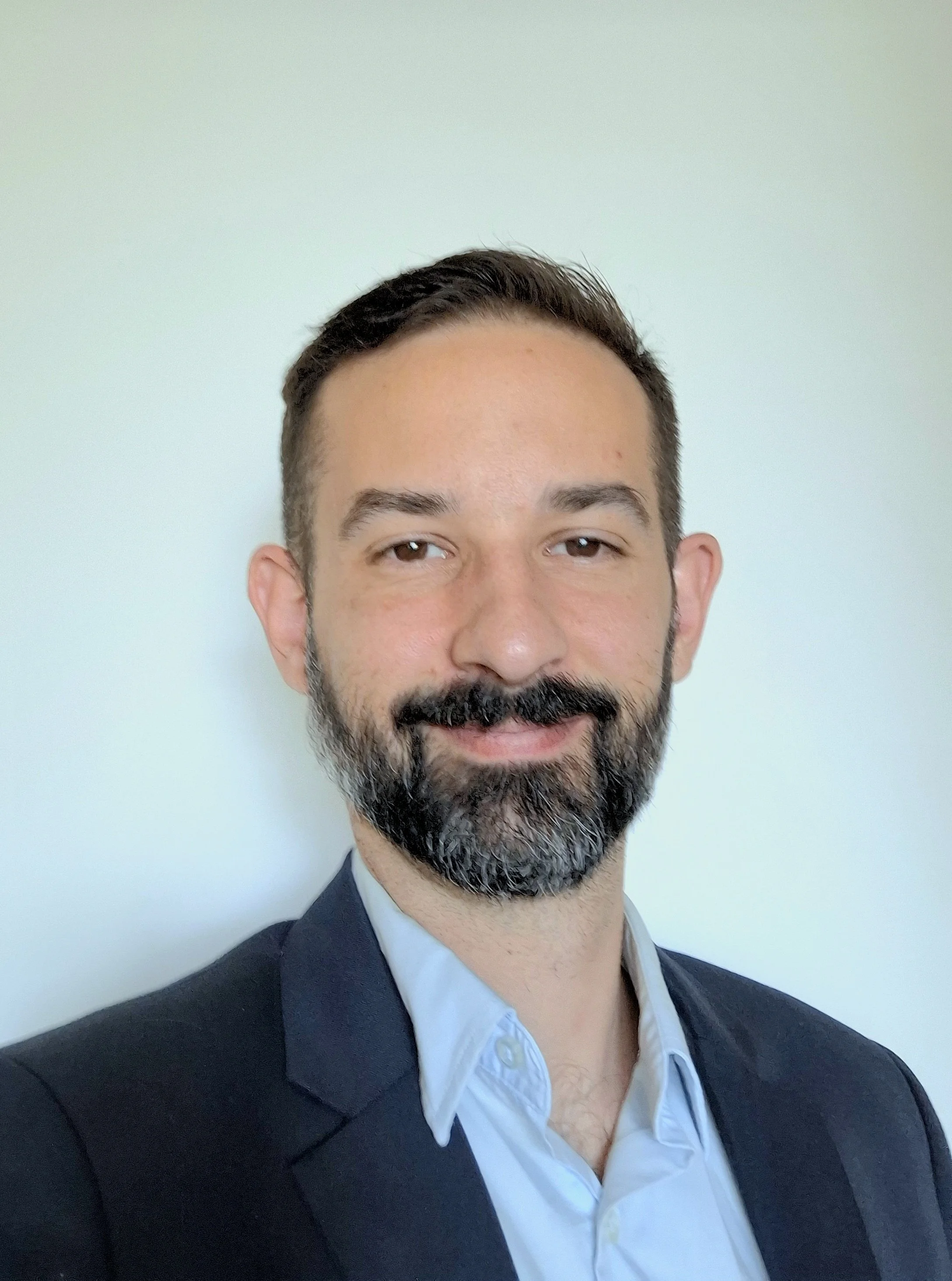 Professional headshot of a man with a beard and short dark hair, wearing a dark suit jacket and a light-colored dress shirt, against a plain light background.