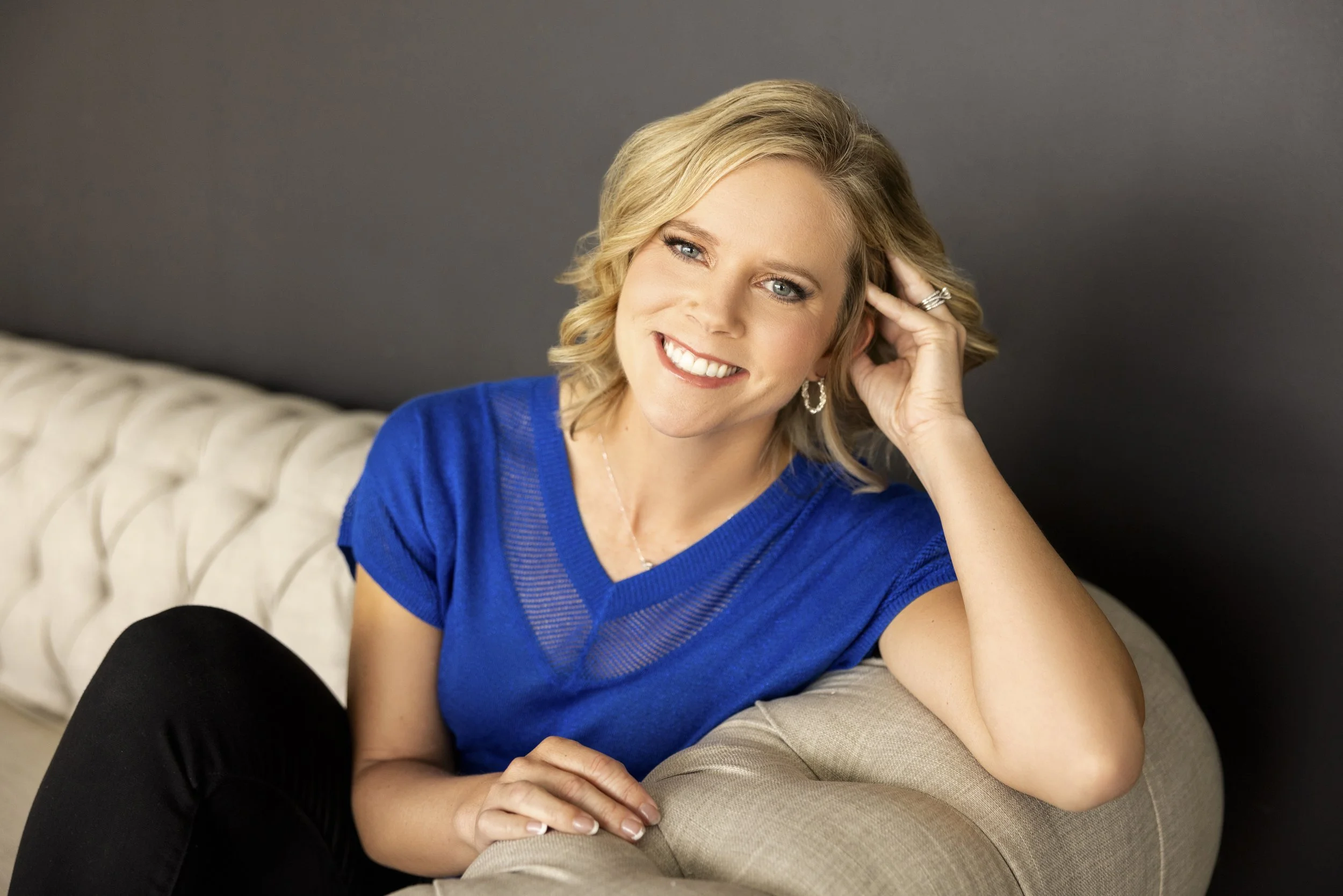 Amy Racki with Racki Consulting, a leadership coaching firm, wearing a blue top, sitting on a beige couch with a dark gray wall background.