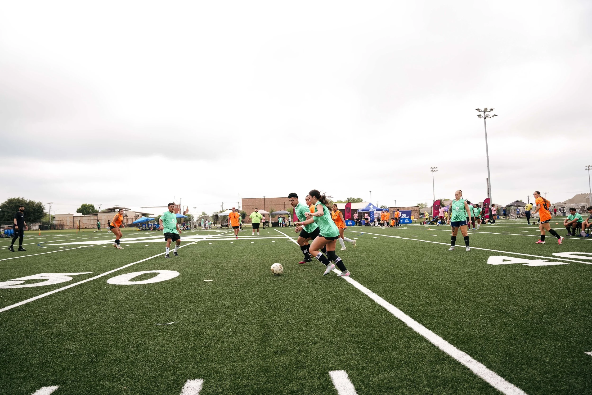 Soccer players practicing on a turf field during a cloudy day, with tents and spectators in the background.