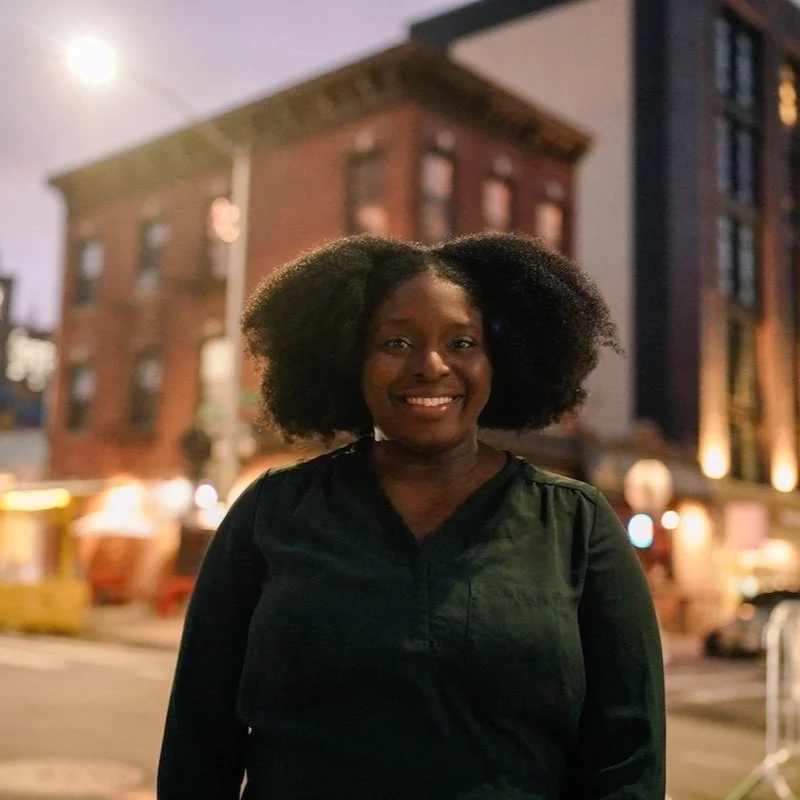 NYC Footy's Zimanta Brissett. A person smiling while standing on a city street in the evening, with a brick building in the background and streetlights illuminating the scene.