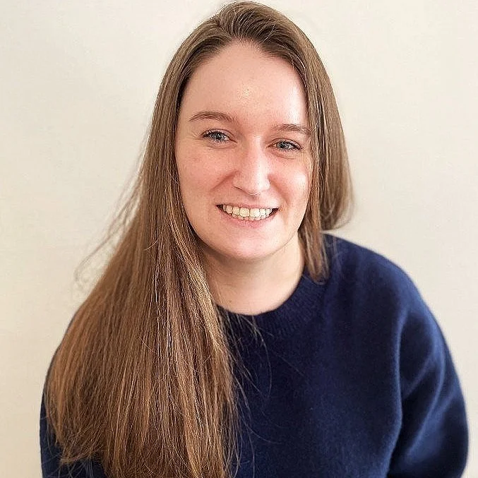 NYC Footy Meghan McGrath. Smiling person with long brown hair wearing a navy sweater against a light background.