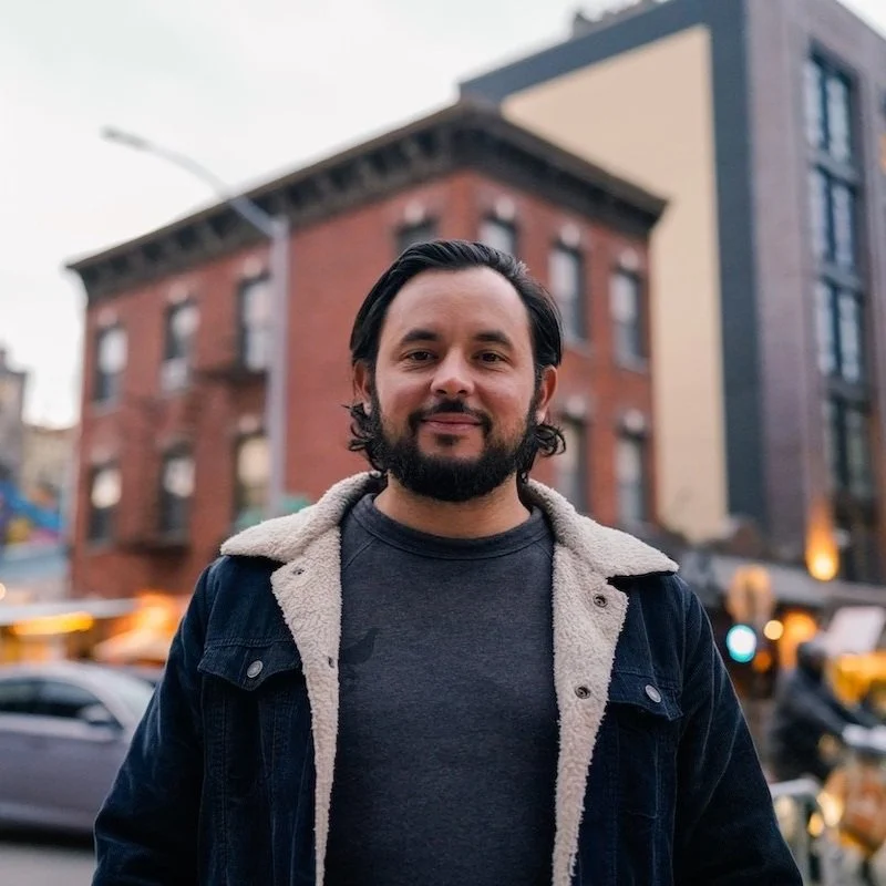 NYC Footy's Gerardo Cueva. Man with beard wearing a denim jacket, standing on a city street, with brick buildings in the background.