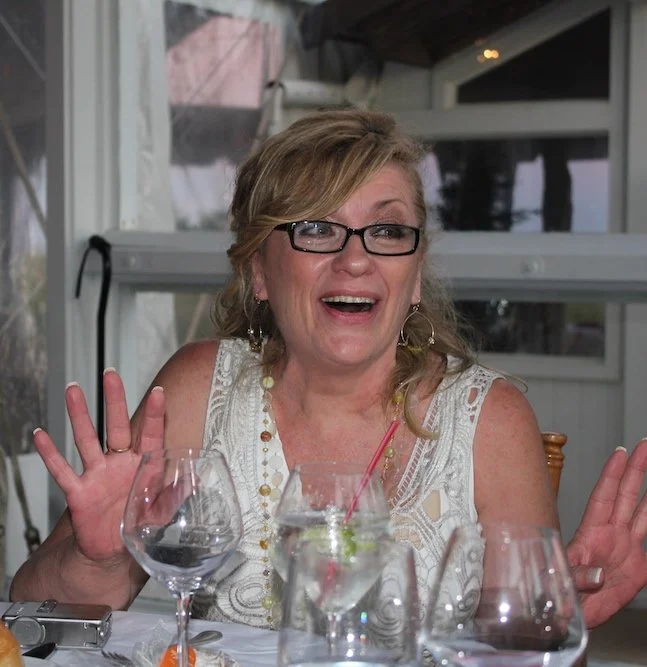 NYC Footy's Kathy Pertew. A woman sitting at a dinner table with several glasses, smiling and gesturing with her hands.