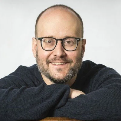 NYC Footy's Billy Lyons. Smiling person with glasses and a beard, wearing a dark sweater, resting chin on hands.