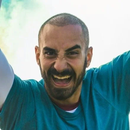 NYC Footy's Gabriel Neves. Man with a shaved head and beard smiling energetically, wearing a blue shirt, with arms raised in excitement.