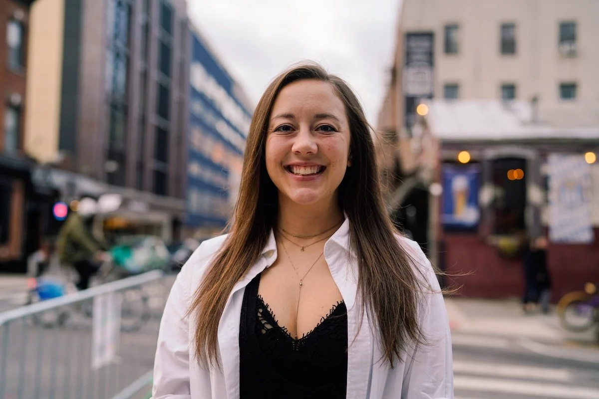 NYC Footy's Kristin Dickerson. A smiling woman in a white shirt standing on a street with blurred buildings in the background.