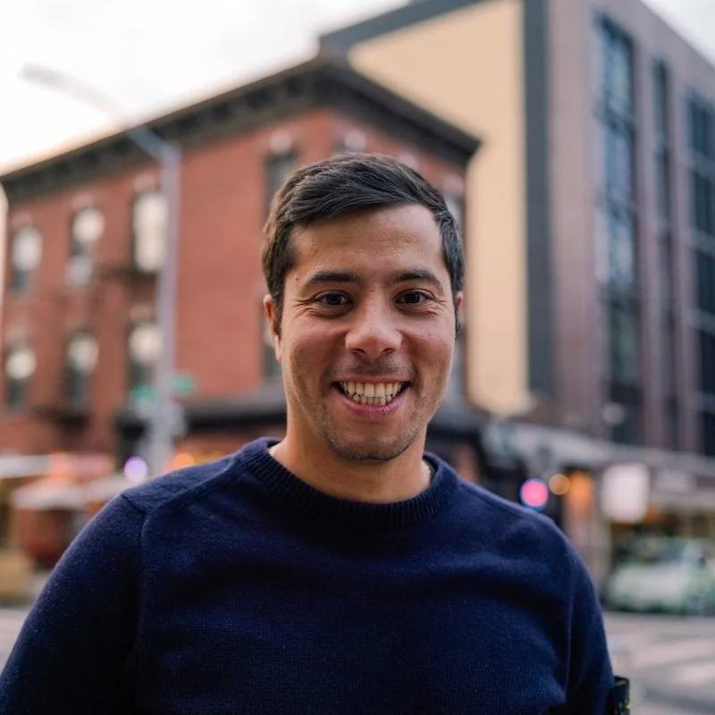 NYC Footy's Gianluca Palanca. A smiling man wearing a dark blue sweater is standing on a city street with brick buildings in the background.