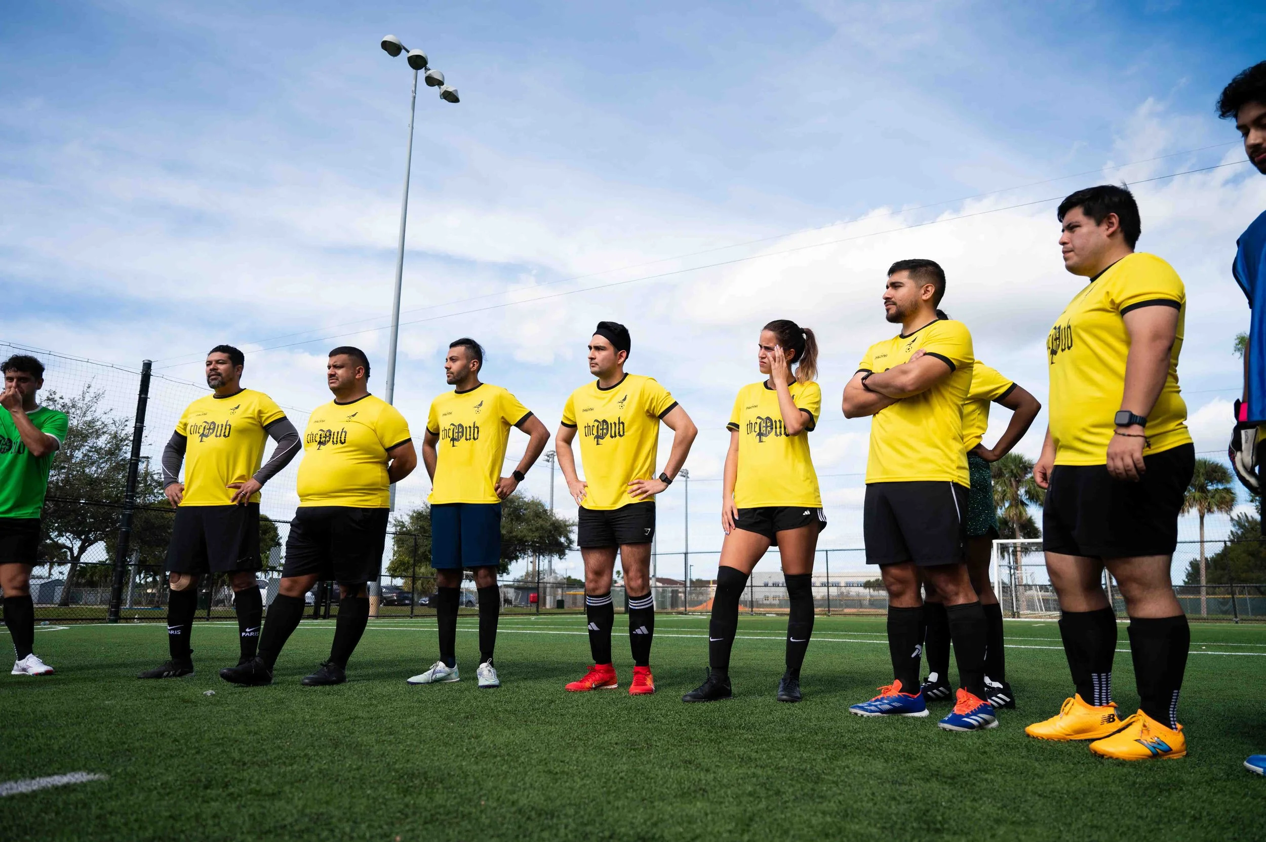 Soccer players standing on a field, some with hands on hips or crossed, wearing yellow and green jerseys, under a partly cloudy sky.